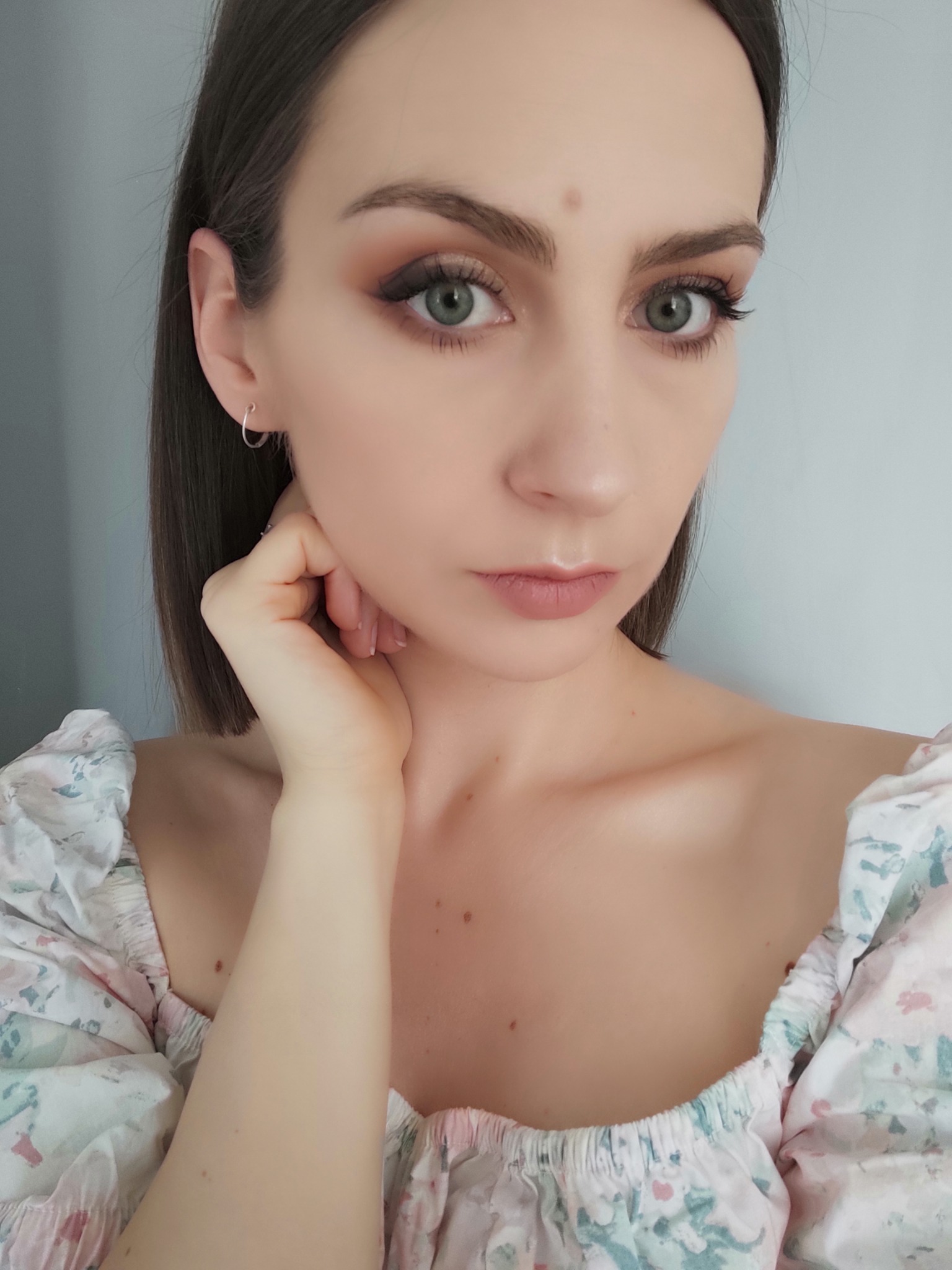 Portret kobiety z delikatnym makijażem oczu w odcieniach brązu, srebrne kolczyki kółka i bluzka w kwiatowy wzór.