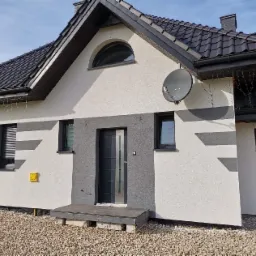 Dwukolorowa elewacja domu jednorodzinnego z szarymi akcentami, widoczna antena satelitarna, okno w kształcie półkola, oraz fragment rusztowania z pracownikiem w tle.