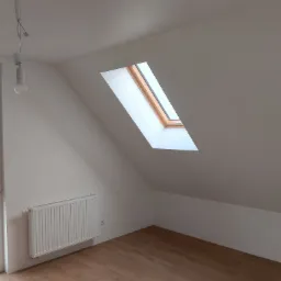 Poddasze po remoncie z drewnianą podłogą, białymi ścianami, oknem dachowym i grzejnikiem, widoczna żarówka na kablu.
