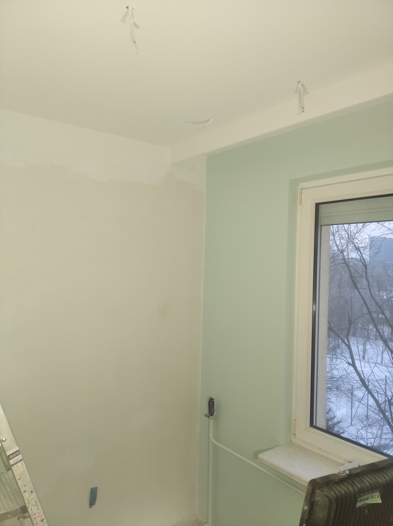 Pokój w trakcie prac wykończeniowych: ściany częściowo pomalowane na biało i miętowo, widoczne przewody wystające z sufitu, fragment drabiny i lampy halogenowej.