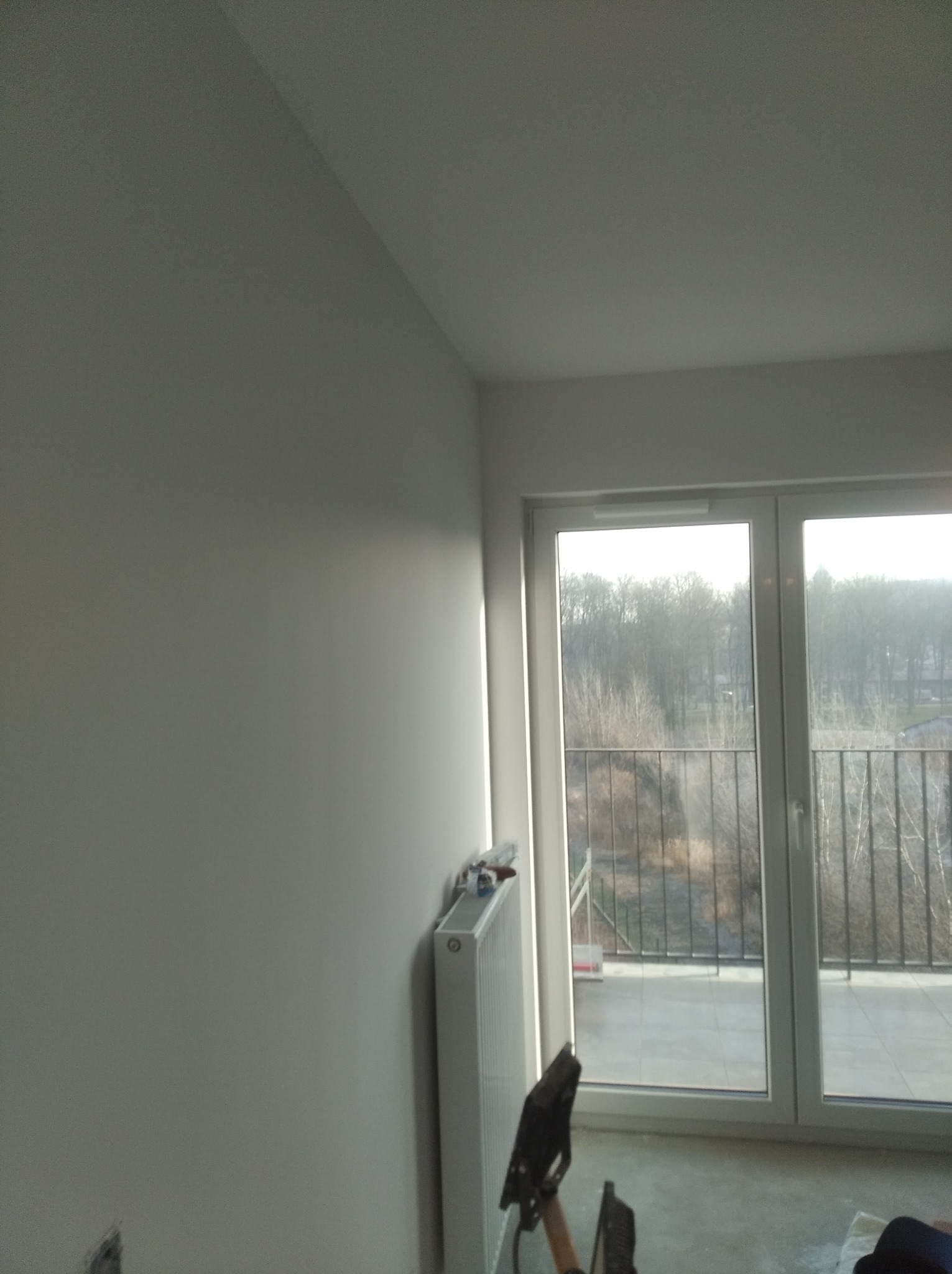 Świeżo pomalowane, jasnoszare ściany w pokoju z balkonem, widoczny grzejnik i narzędzia malarskie w dolnej części kadru.