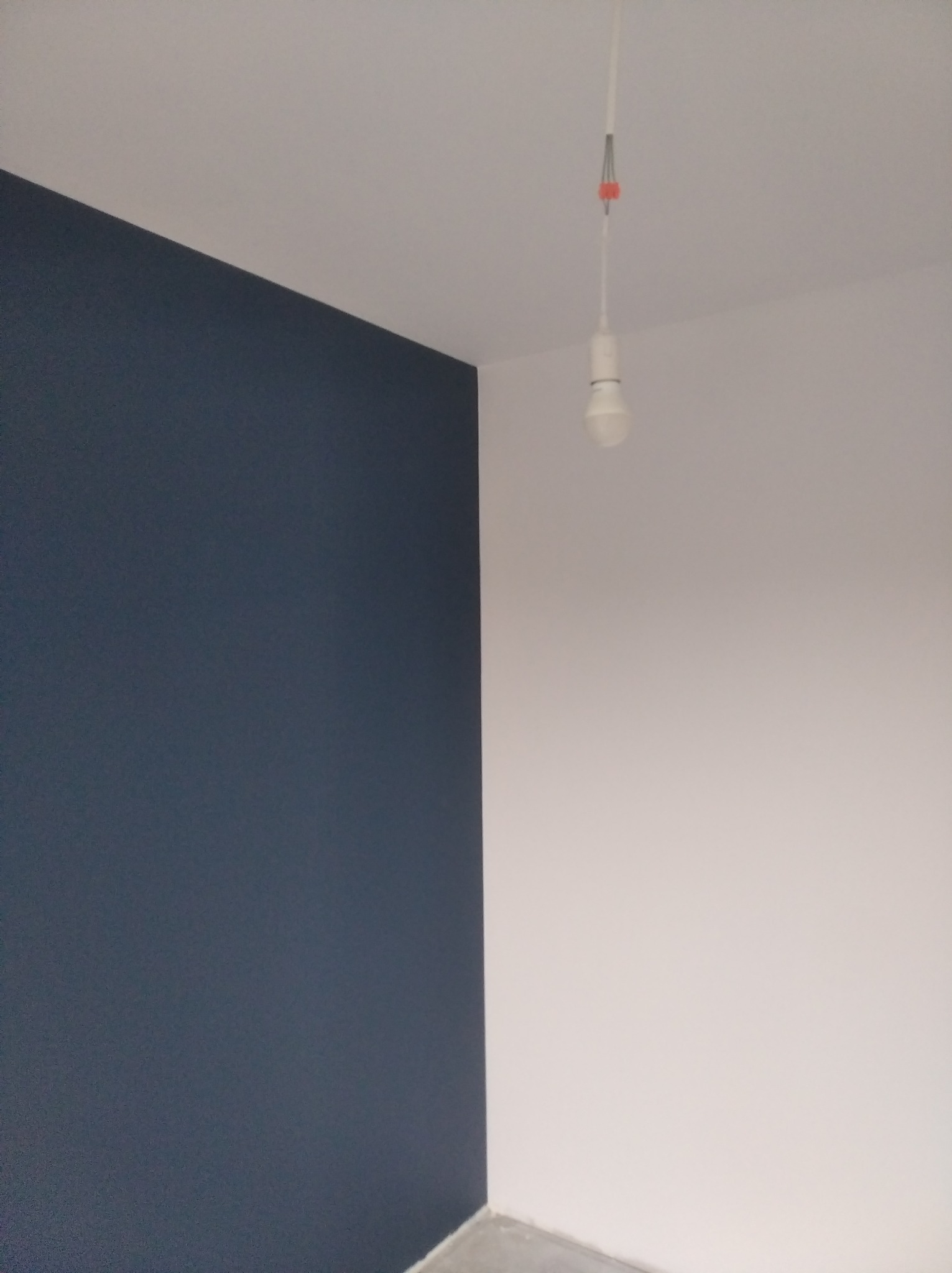 Świeżo pomalowane wnętrze pokoju z jedną ścianą w kolorze granatowym, a drugą białą, pod sufitem wisząca żarówka na kablu.