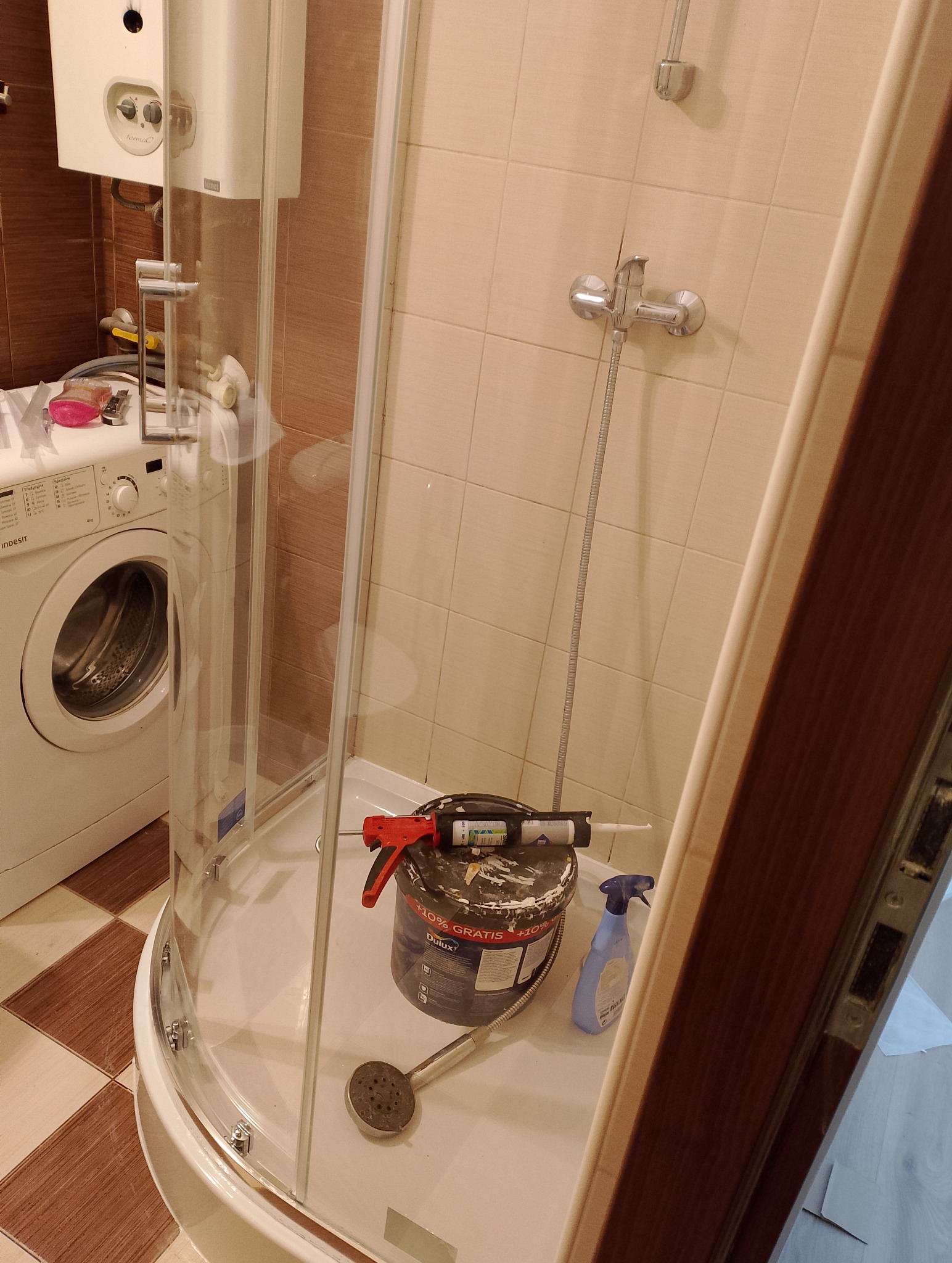 Wnętrze łazienki w trakcie remontu, widoczna kabina prysznicowa z wiadrem farby Dulux, pistoletem do silikonu, spryskiwaczem i słuchawką prysznicową wewnątrz, obok pralka.