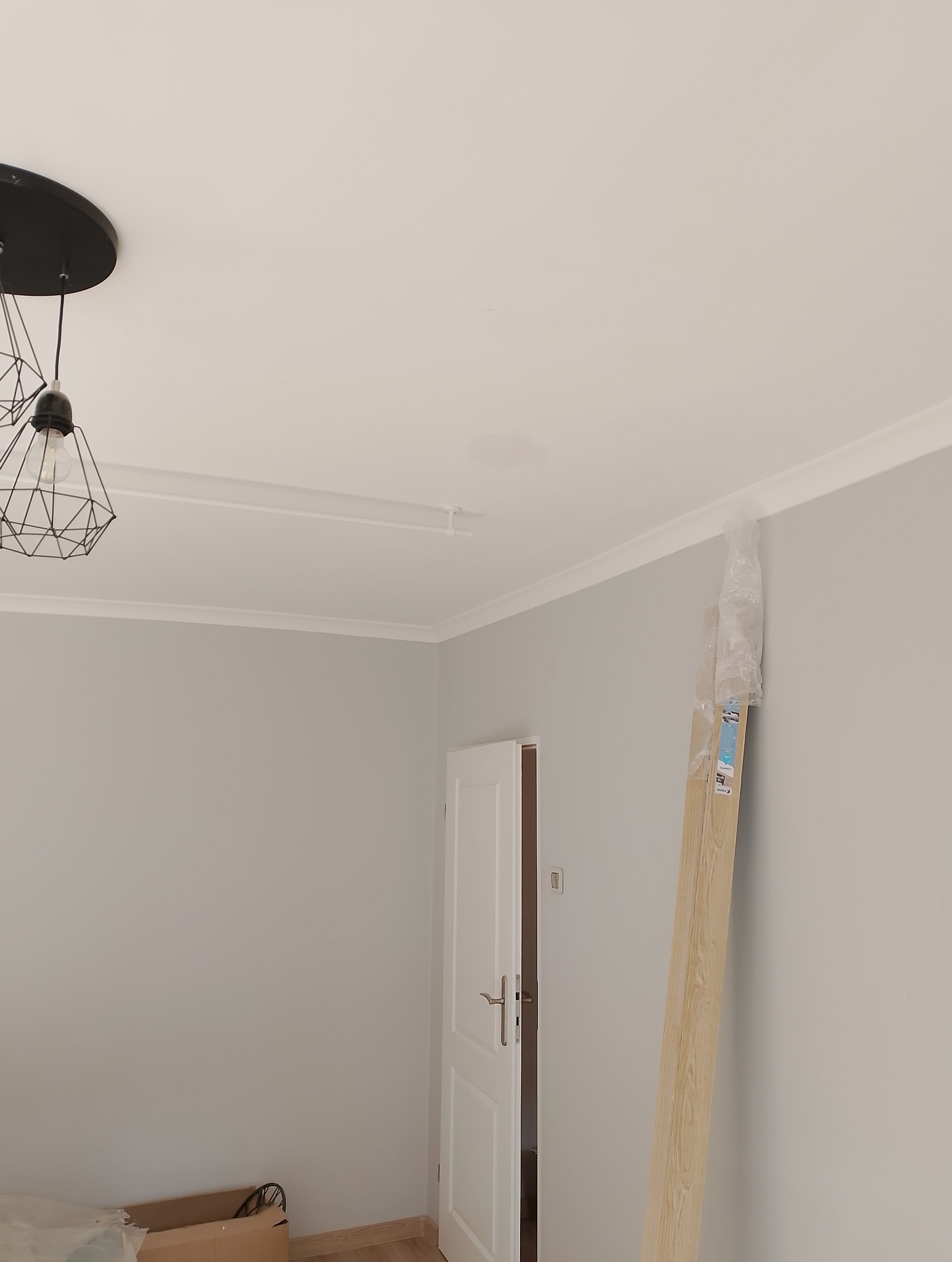 Pokój po remoncie: świeżo pomalowane szare ściany, biały sufit z listwami dekoracyjnymi, nowoczesna lampa wisząca, drzwi z ościeżnicą i panele podłogowe w folii oparty o ścianę.