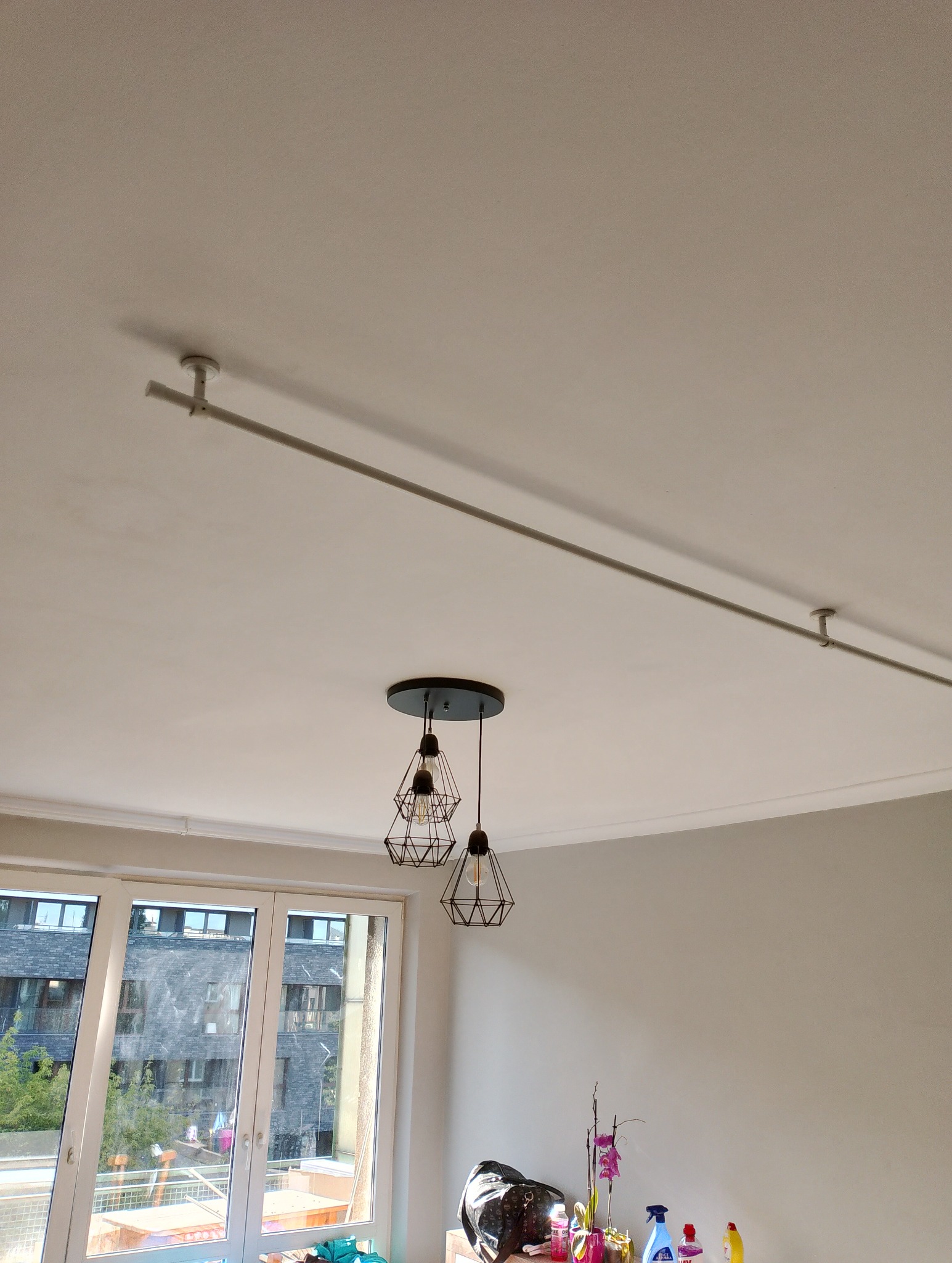 Biały sufit z zamontowaną szyną oświetleniową i wiszącą lampą z trzema geometrycznymi kloszami, widok z dołu na tle okna i fragmentu ściany.