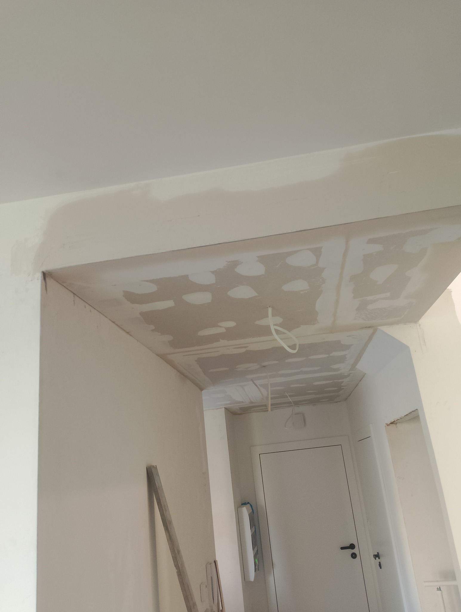 Sufit w trakcie szpachlowania z widocznymi punktami po wkrętach, wystające przewody oświetleniowe, fragment ściany i drzwi w tle