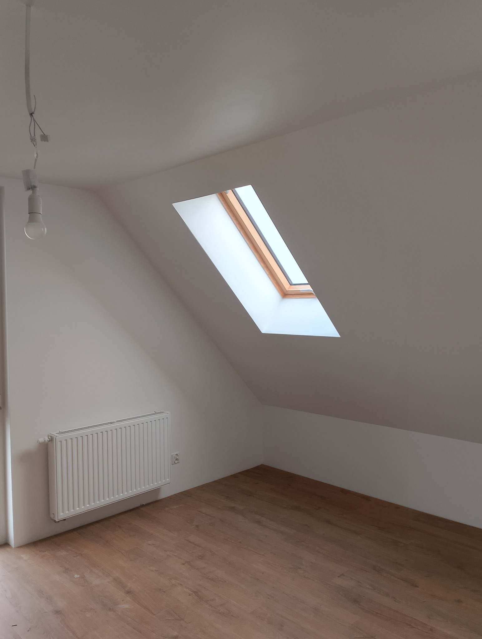 Poddasze po remoncie z drewnianą podłogą, białymi ścianami, oknem dachowym i grzejnikiem, widoczna żarówka na kablu.