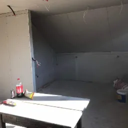 Poddasze w trakcie adaptacji: widoczne ściany wykończone płytami kartonowo-gipsowymi, wystające przewody elektryczne, na pierwszym planie stół z napojami i przekąskami.