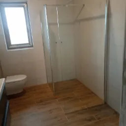 Nowoczesna kabina prysznicowa z transparentnego szkła w wyremontowanej łazience z drewnopodobną podłogą, widoczna toaleta i okno z naturalnym światłem.