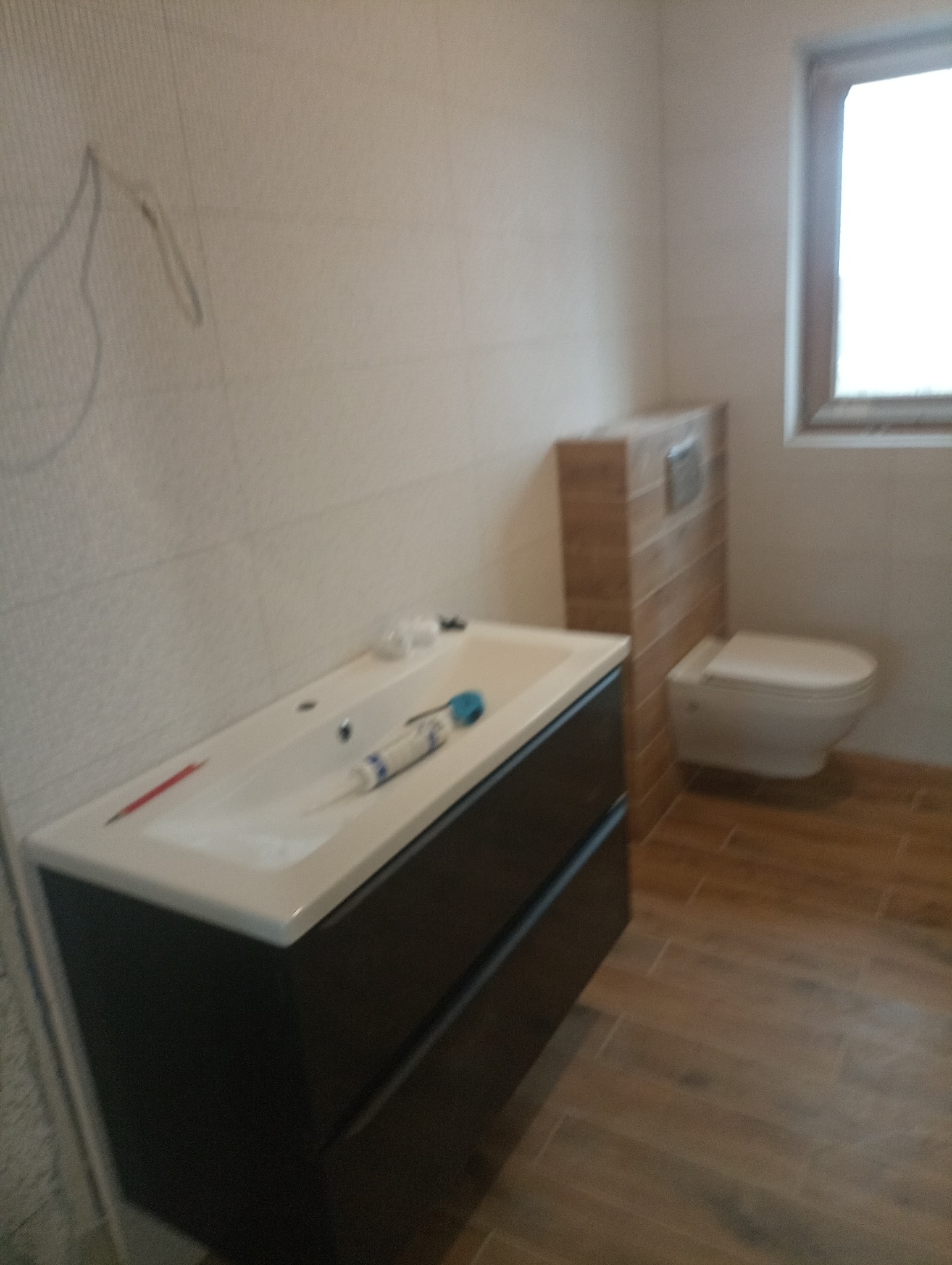 Montaż umywalki z szafką w łazience, widoczna biała umywalka z czarną szafką, silikon sanitarny, płytki podłogowe imitujące drewno i podwieszana toaleta z zabudową.