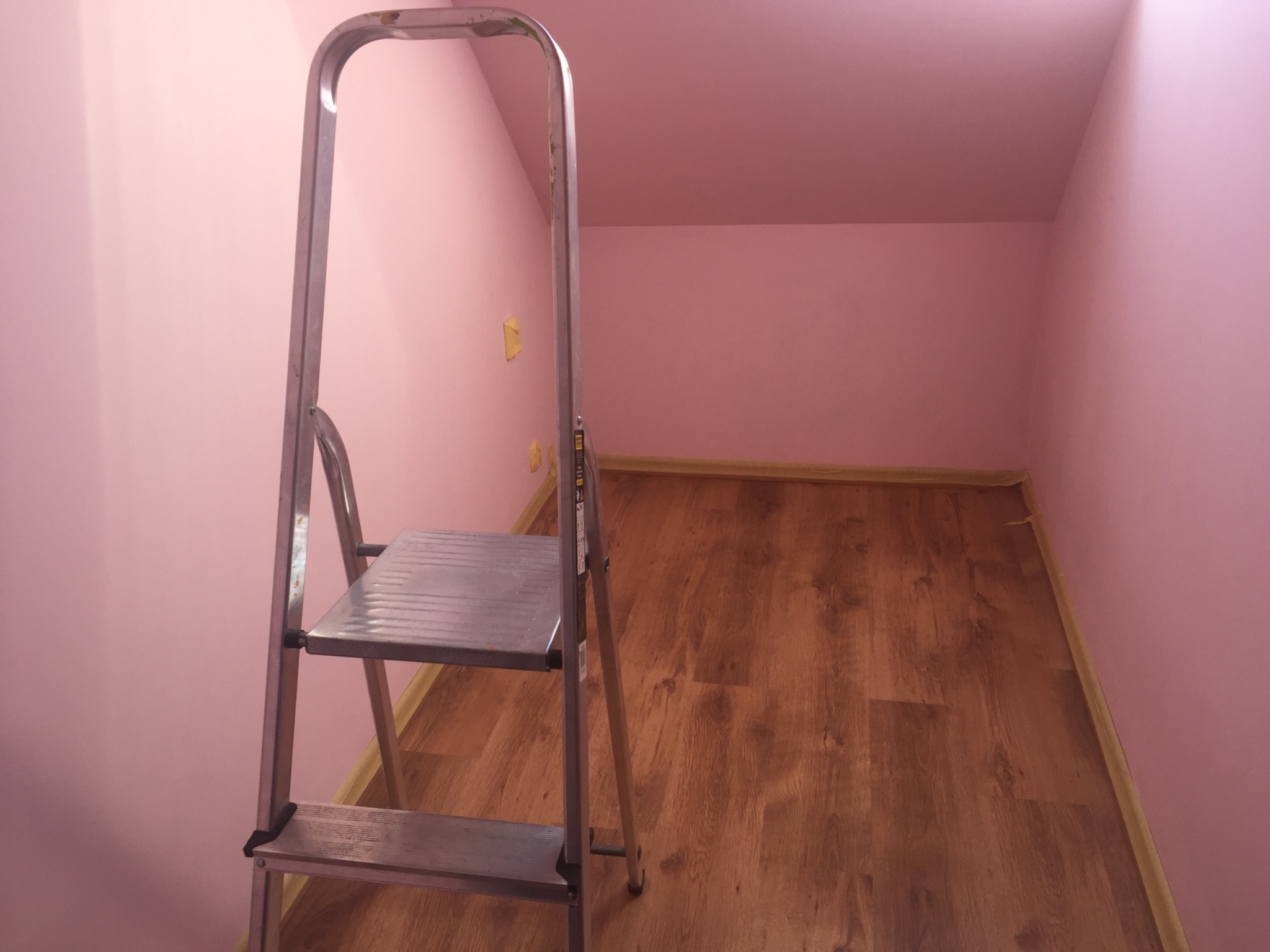 Metalowa drabina stojąca w rogu pomalowanego na różowo pokoju na drewnianej podłodze, z żółtą taśmą malarską widoczną na ścianie.