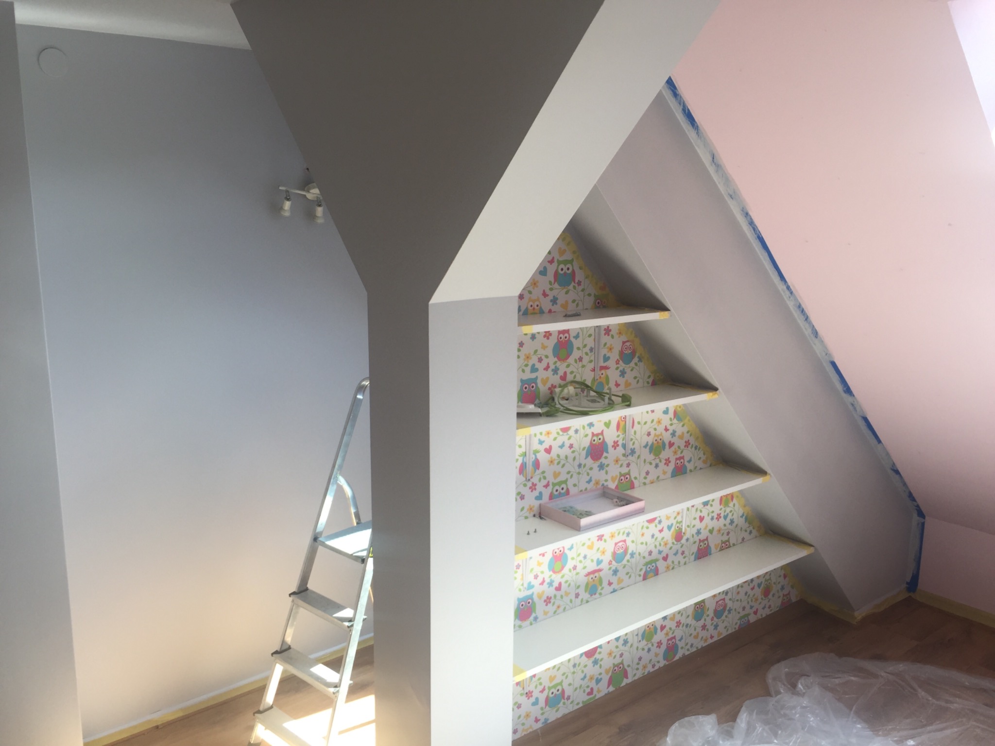 Pokój na poddaszu w trakcie prac: pomalowana szara ściana, różowa ściana zabezpieczona taśmą malarską, białe półki w skosie z tapetą w sowy, drabina malarska.
