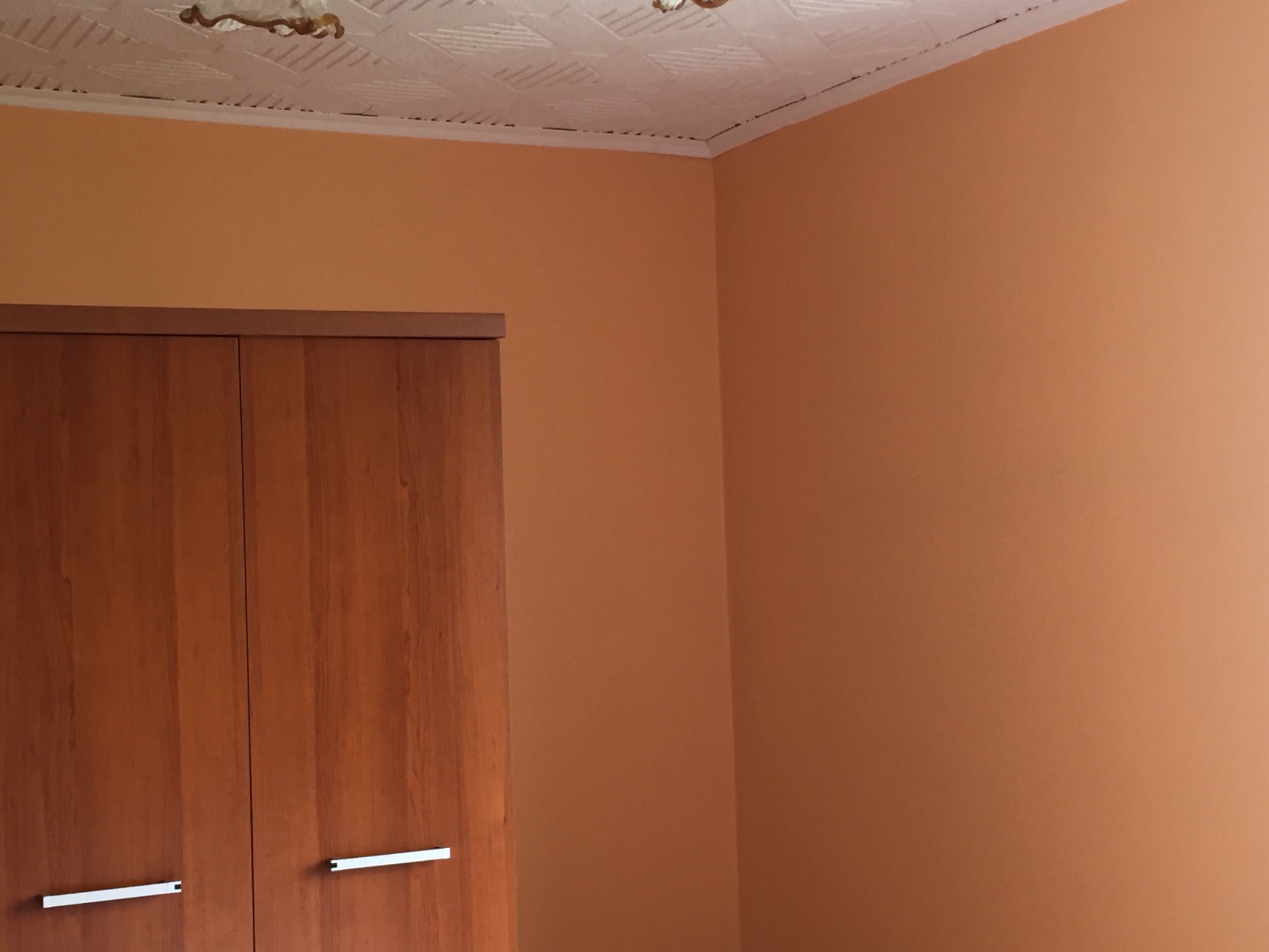 Pomarańczowe ściany w pokoju po odświeżeniu, widoczna szafa z jasnego drewna i biały sufit kasetonowy z delikatnym wzorem.