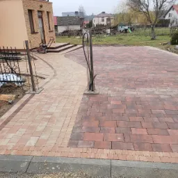 Ułożona kostka brukowa w dwóch odcieniach czerwieni i beżu tworząca wzór przed domem z widoczną ubijarką w tle, obramowana szarą kostką brzegową.