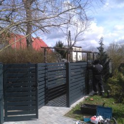 NOSTAL ŚLUSARSTWO Rafał Nosalski - Montaż nowoczesnego, poziomego ogrodzenia z metalowych profili w kolorze grafitowym; widoczny pracownik w kombinezonie podczas instalacji przy użyciu narzędzi na tle domu z czerwoną dachówką.