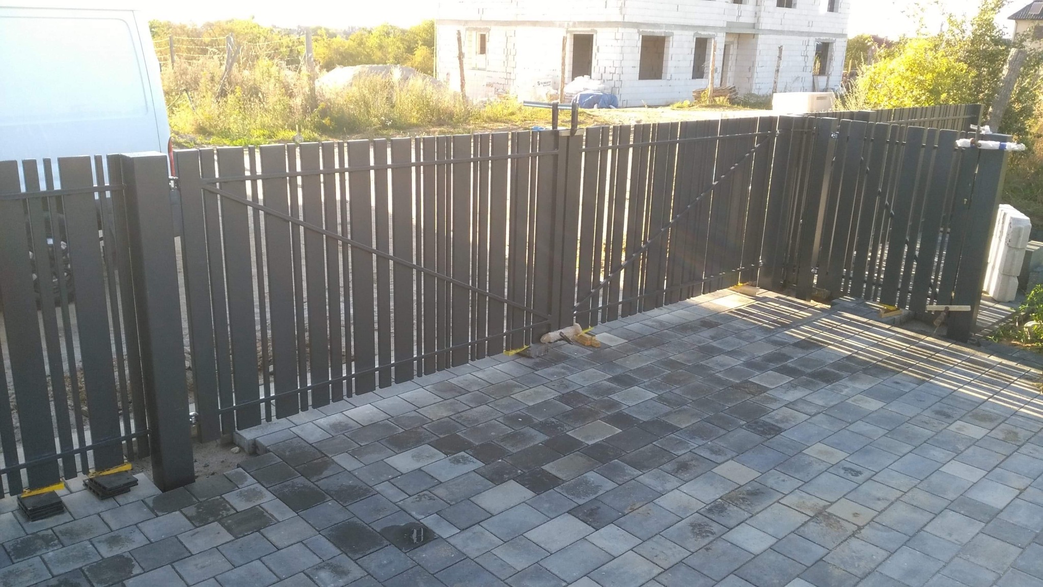 Ciemnoszare, pionowe przęsła ogrodzenia metalowego w trakcie montażu; widoczne żółte podkładki poziomujące, kostka brukowa i budynek w stanie surowym w tle.