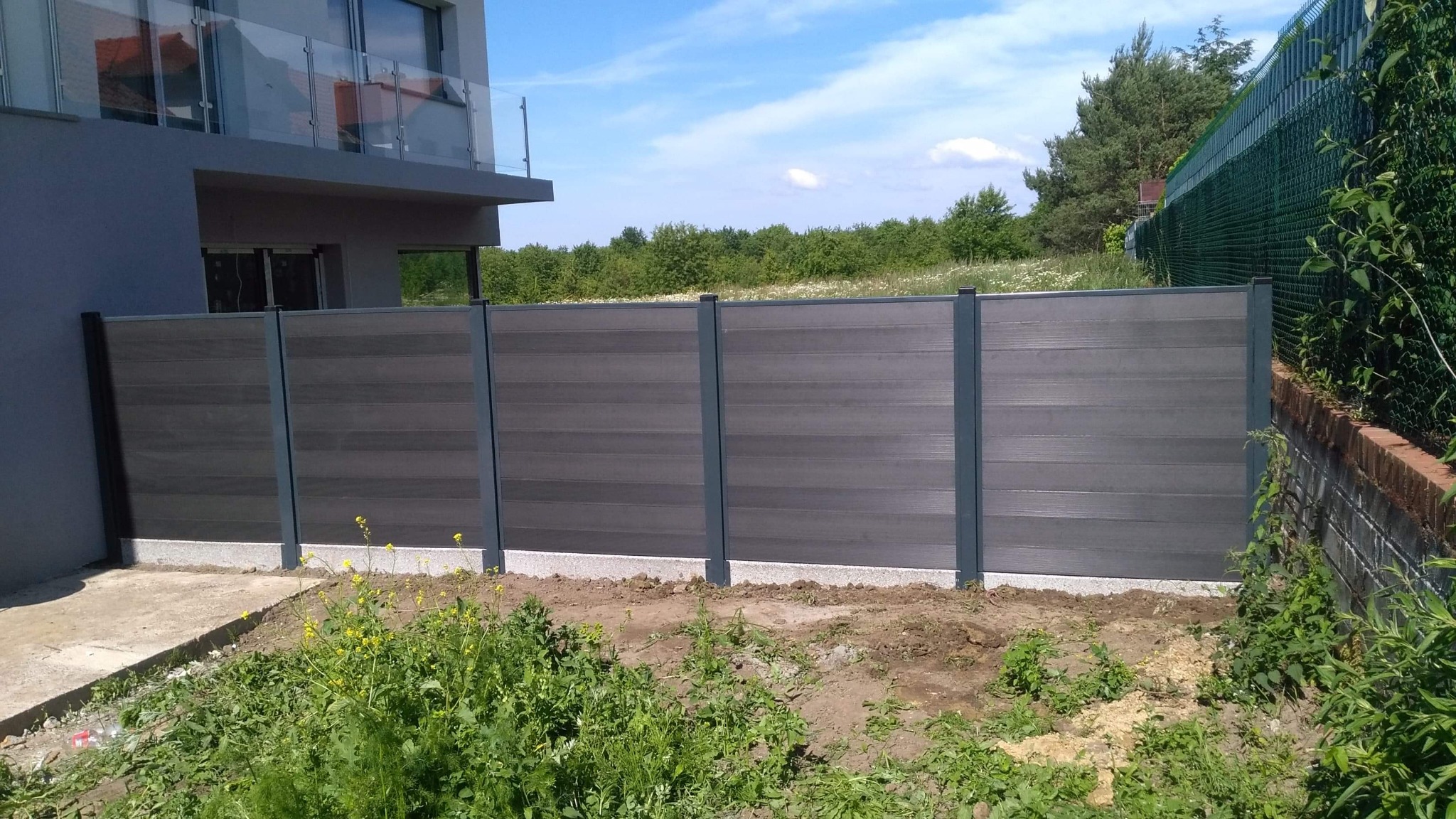 Nowoczesne ogrodzenie z poziomych paneli kompozytowych w kolorze grafitowym, z widocznymi stalowymi słupkami, na tle domu z balkonem i zieleni.