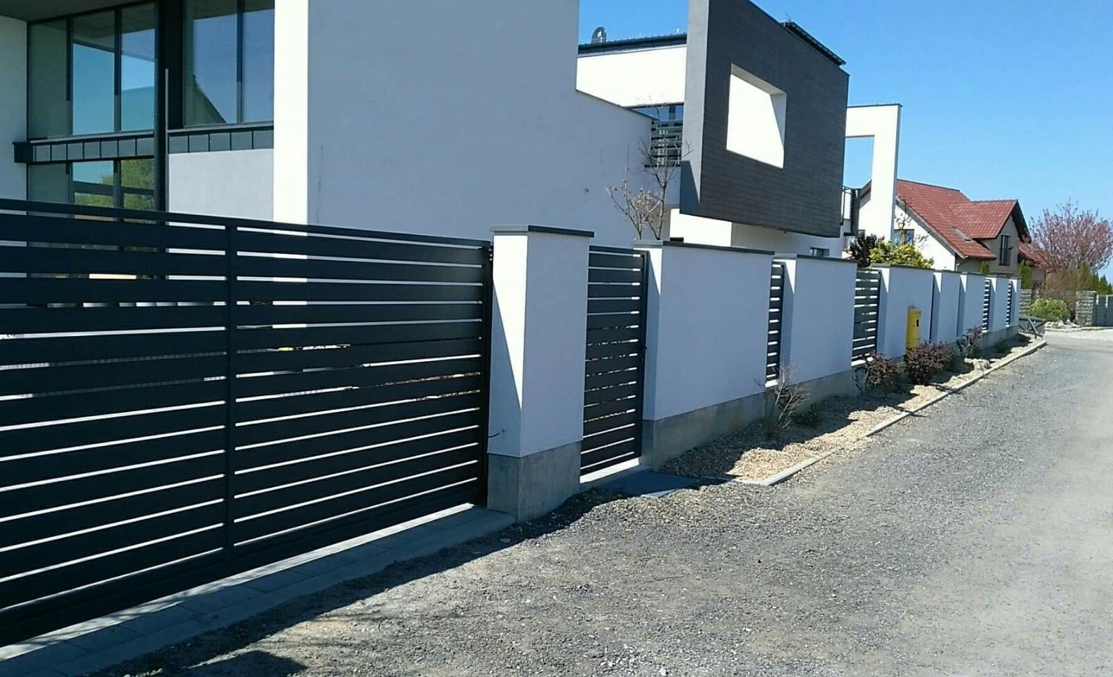 Nowoczesne ogrodzenie z poziomych, czarnych profili metalowych, osadzone na białych, murowanych słupkach z betonową podstawą, na tle minimalistycznego domu z dużymi oknami.