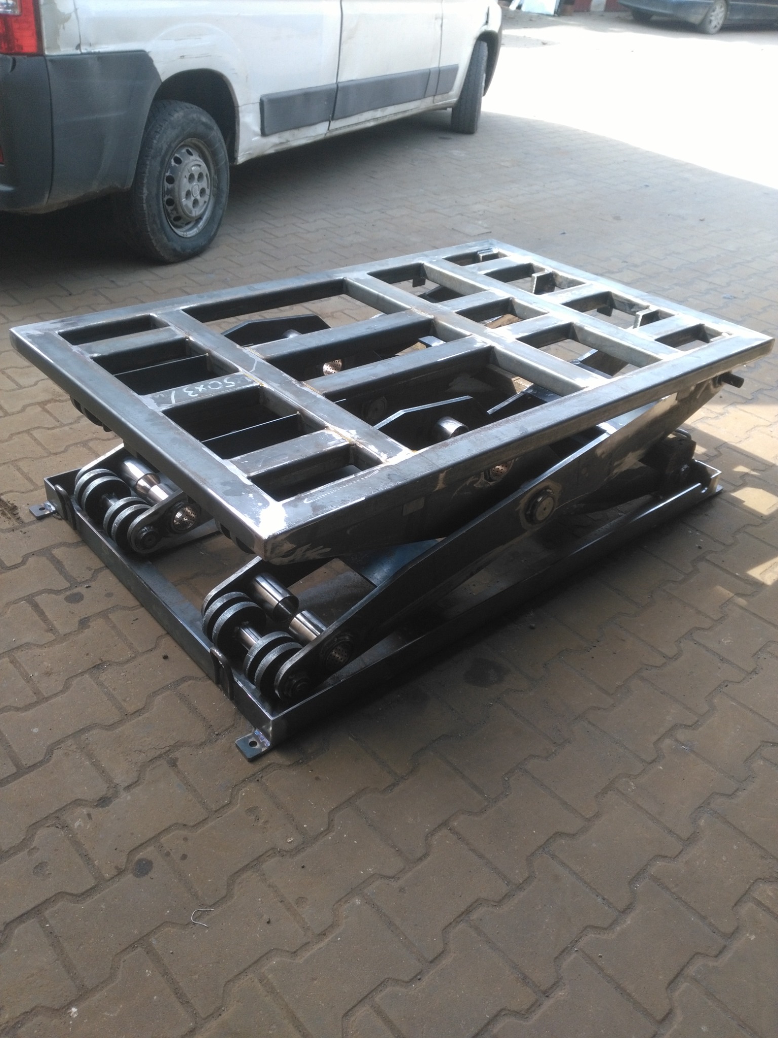 Surowa, metalowa platforma podnośnikowa z kratownicą, widoczna na zewnątrz na tle zaparkowanego pojazdu dostawczego.