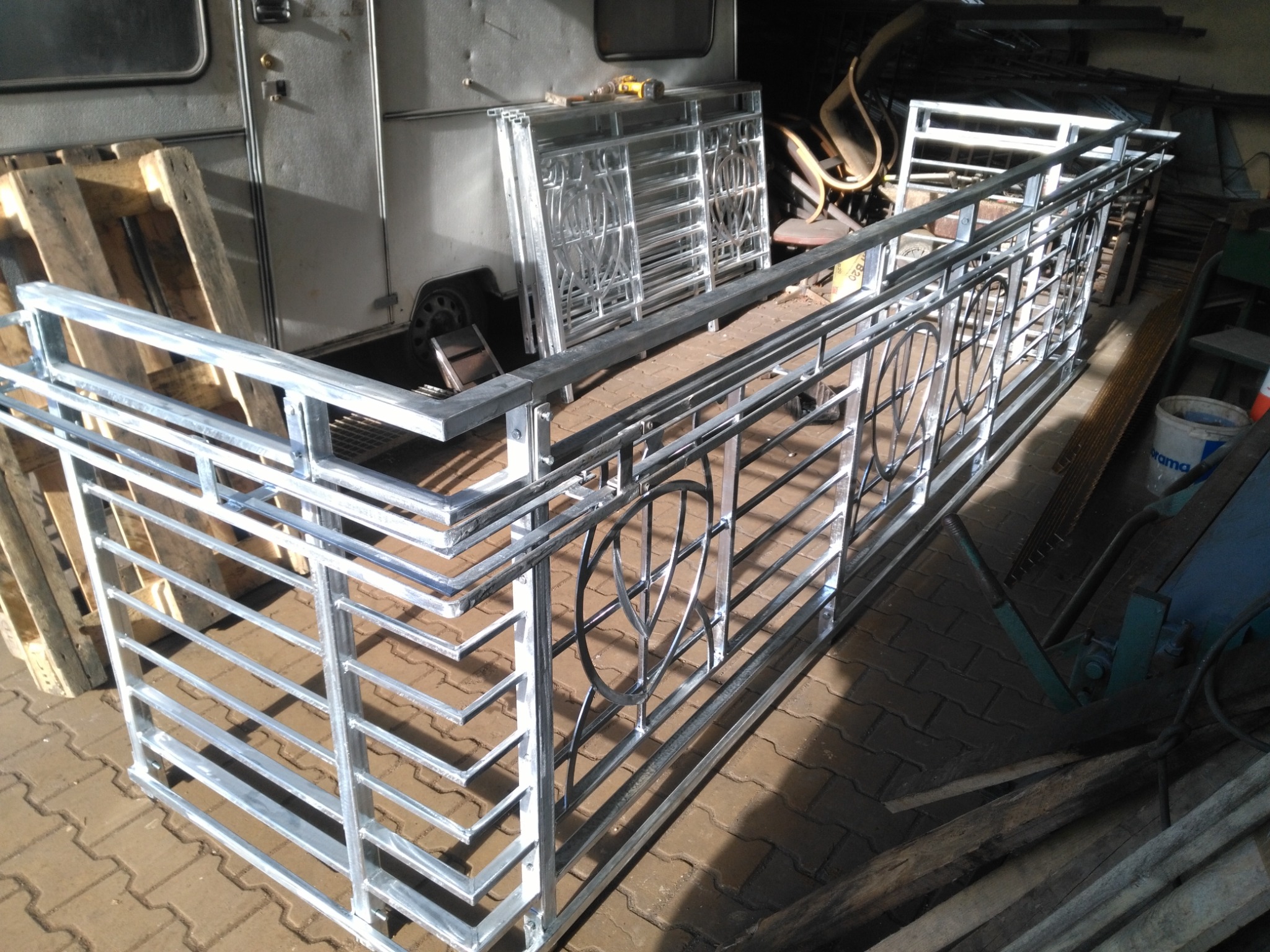 Surowy metalowy fragment balustrady z motywem liścia, przygotowany do montażu, widoczny na tle warsztatu z drewnianymi paletami i przyczepą.