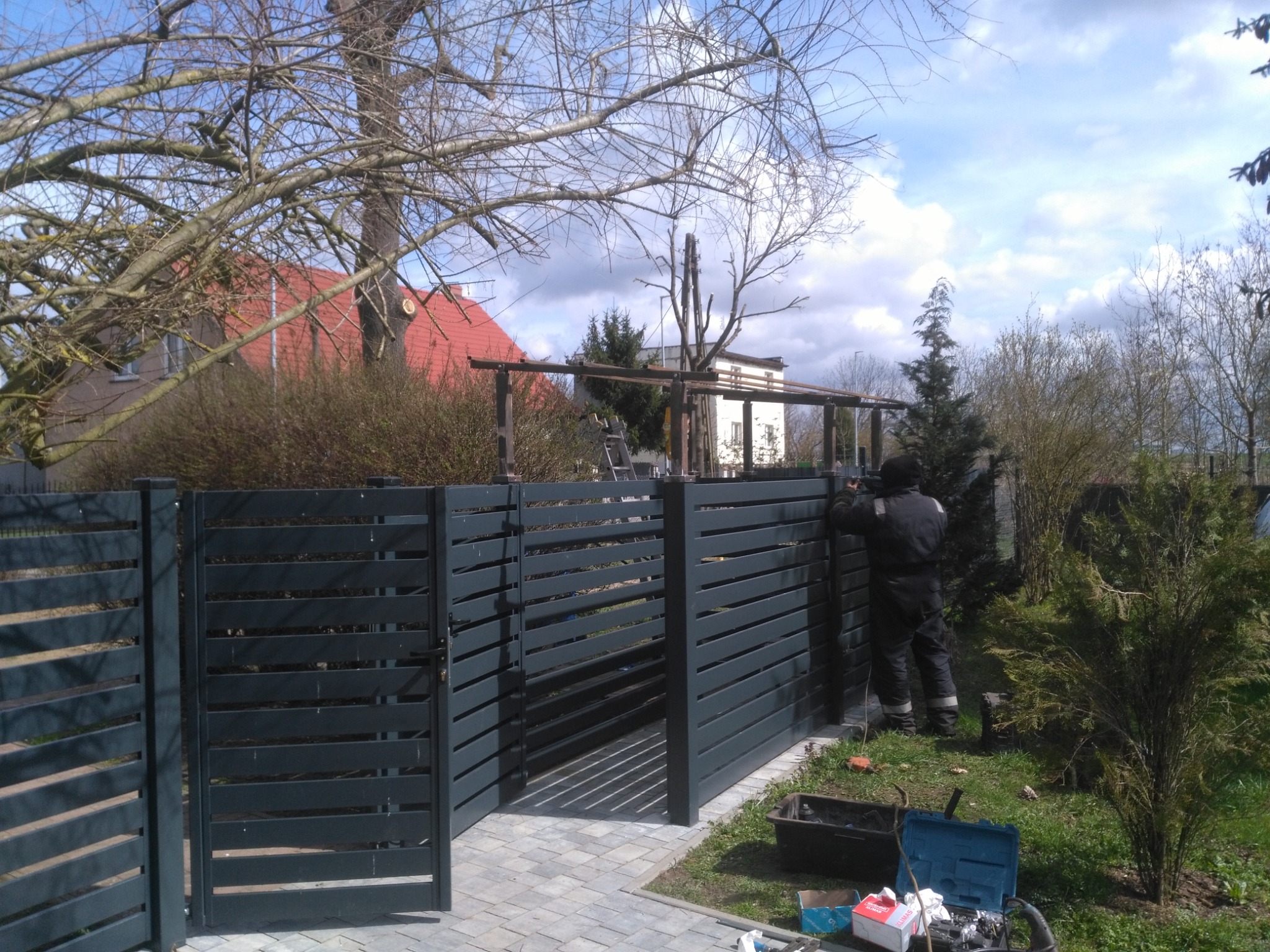 Montaż nowoczesnego, poziomego ogrodzenia z metalowych profili w kolorze grafitowym; widoczny pracownik w kombinezonie podczas instalacji przy użyciu narzędzi na tle domu z czerwoną dachówką.