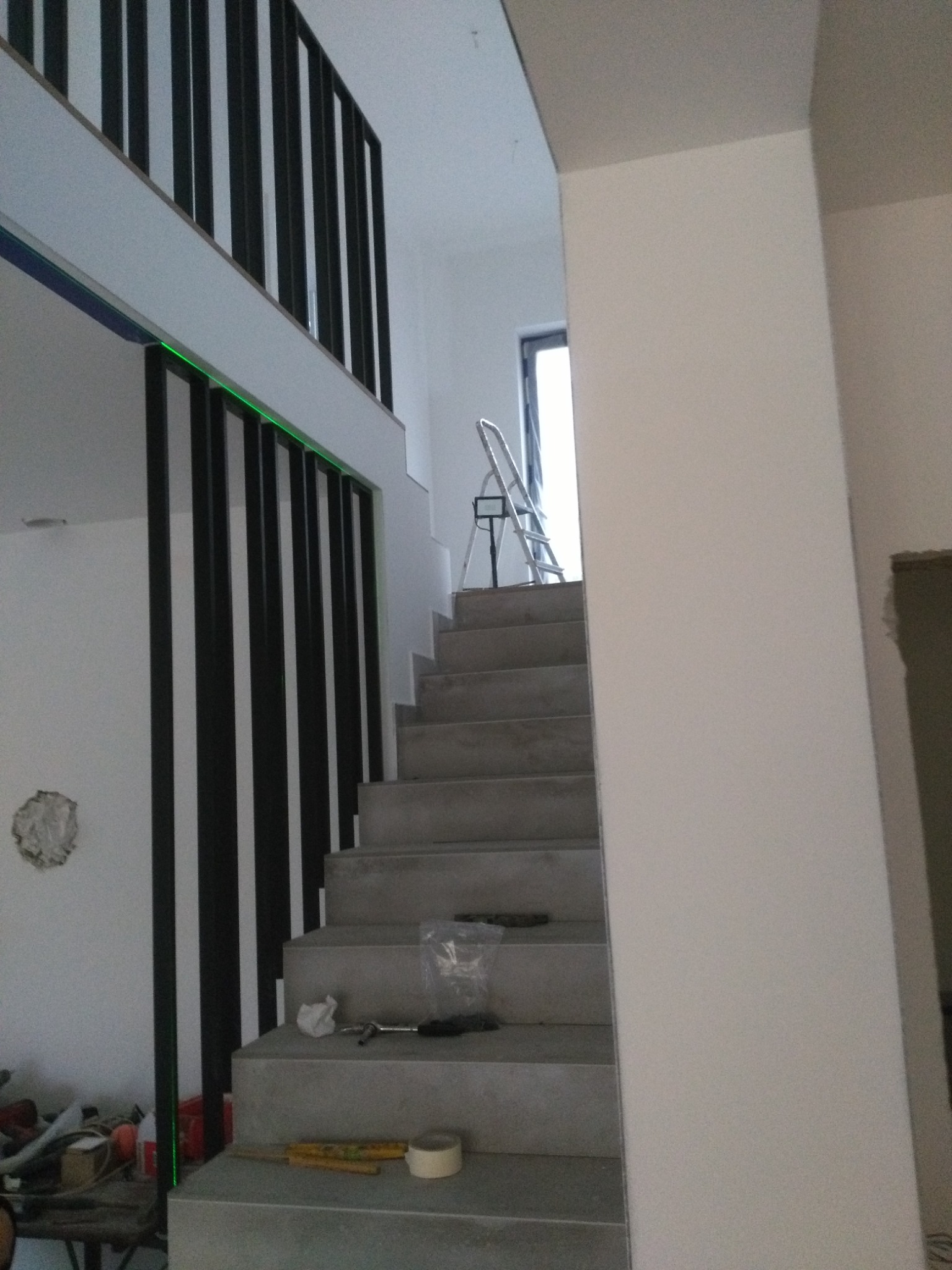 Montaż balustrady z czarnych, metalowych słupków przy schodach z widocznym laserowym poziomowaniem i narzędziami na stopniach.