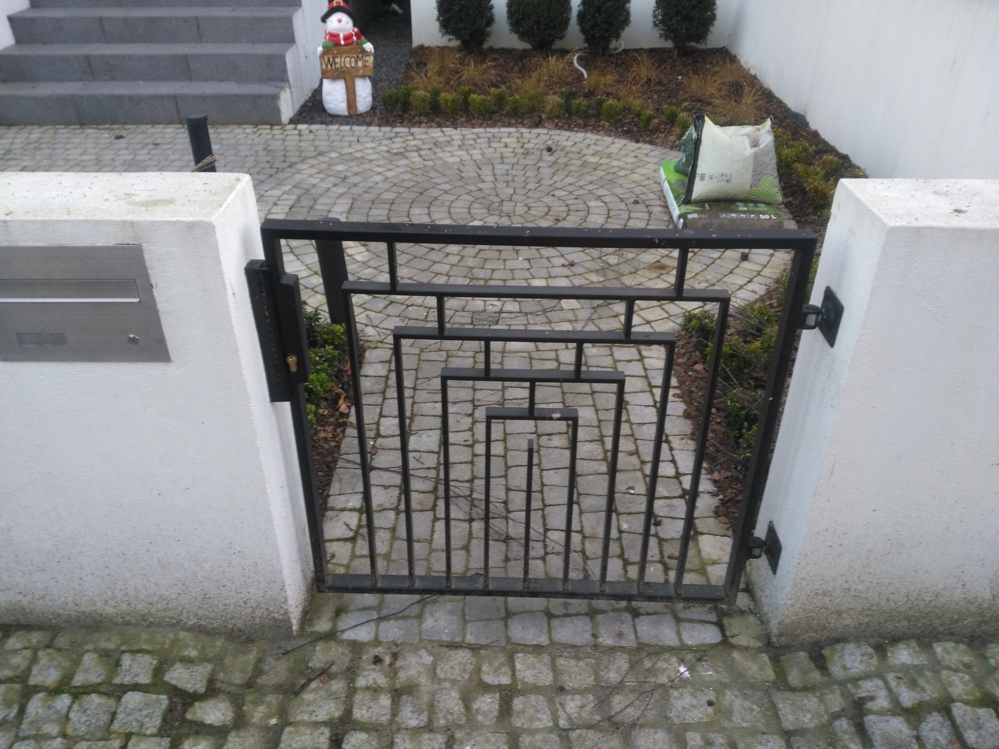 Czarna metalowa furtka o geometrycznym wzorze, osadzona między białymi słupkami na brukowanym podjeździe. W tle widoczny trawnik, krzewy i dekoracja w postaci bałwana.