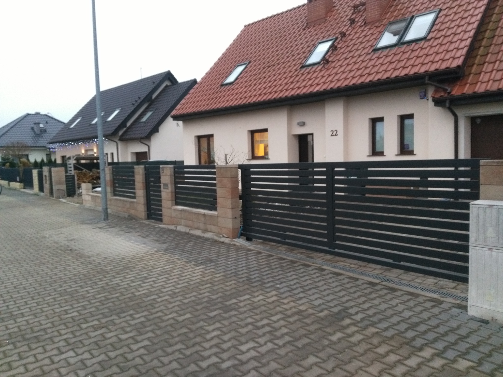 Nowoczesne ogrodzenie z poziomych, metalowych profili w kolorze antracytowym, zamontowane przed domem z czerwoną dachówką i numerem 22, widoczne z poziomu kostki brukowej.