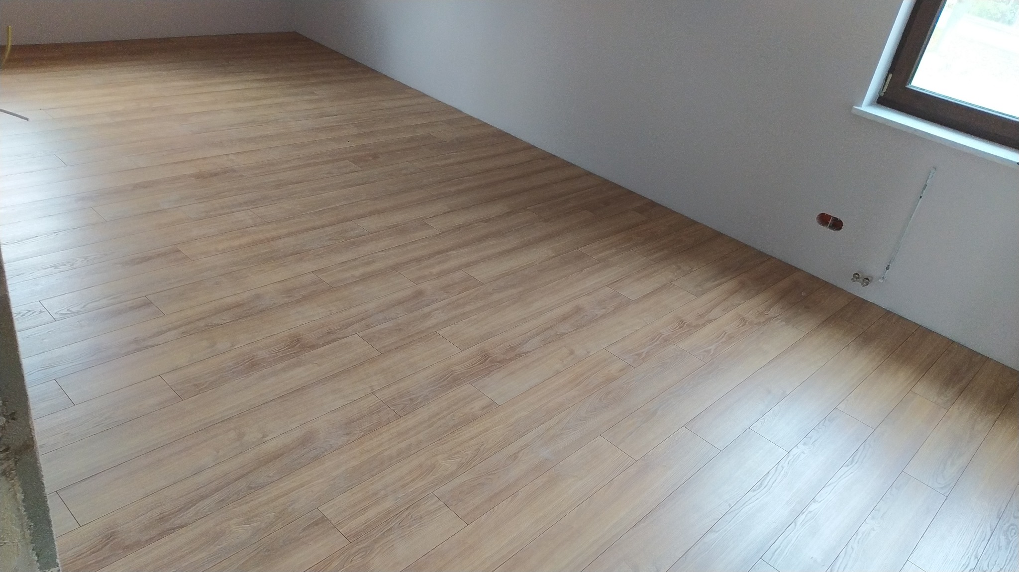 Jasne panele podłogowe ułożone w skos w nowym pomieszczeniu, widoczne gniazdka elektryczne i fragment okna.