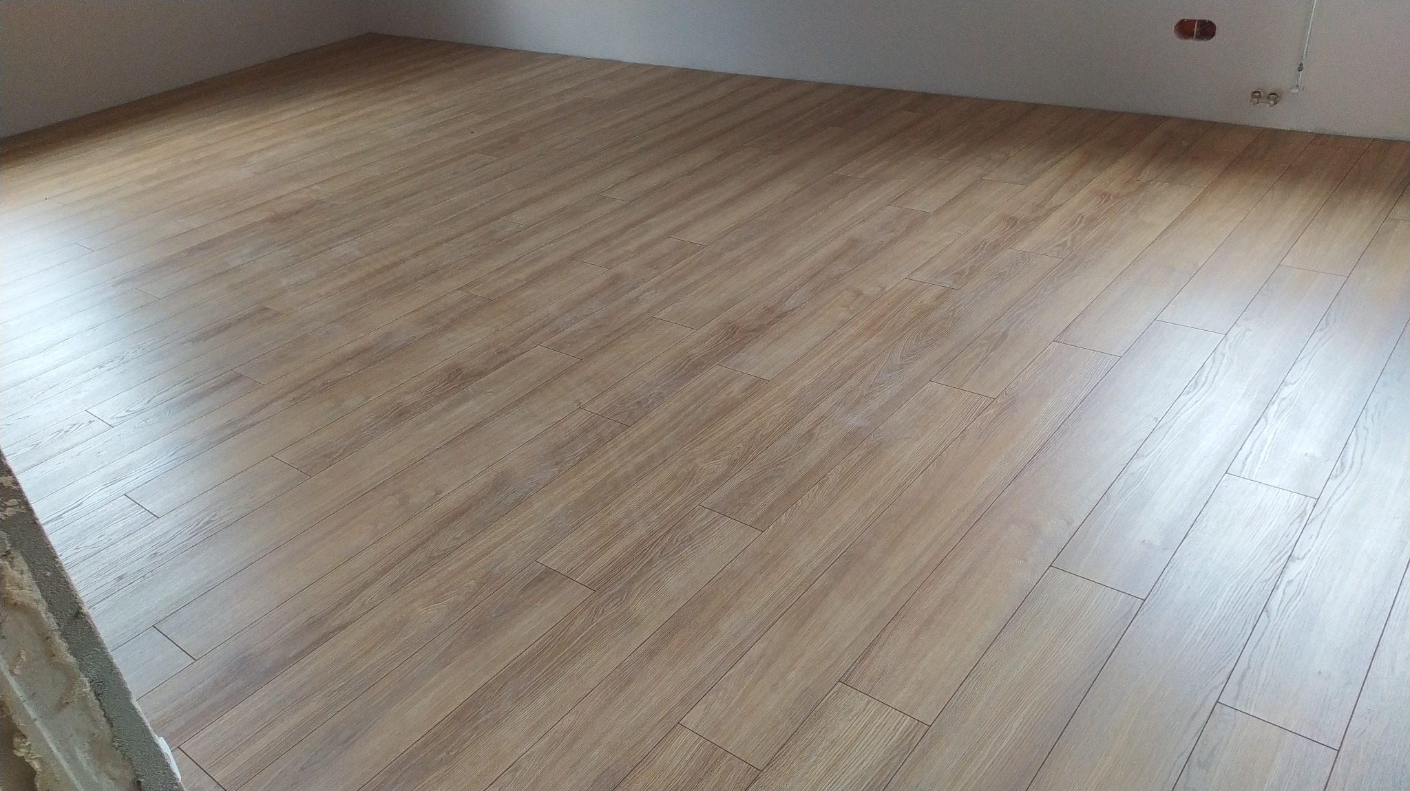 Jasnobrązowe panele podłogowe ułożone w nowym pokoju, widoczne gniazdka elektryczne na ścianie i fragment surowej ściany przy podłodze.