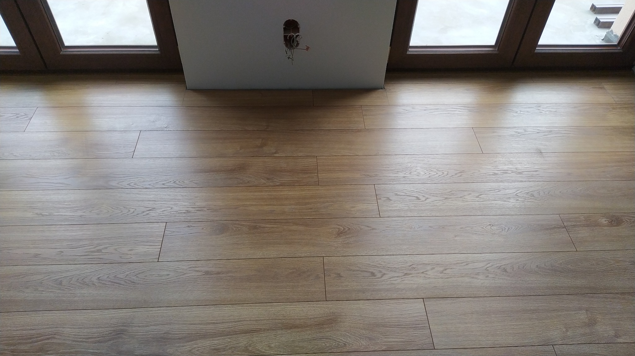 Nowo ułożona podłoga z paneli laminowanych o jasnym odcieniu drewna, widoczna ściana z wystającymi przewodami elektrycznymi i fragment ramy okiennej.