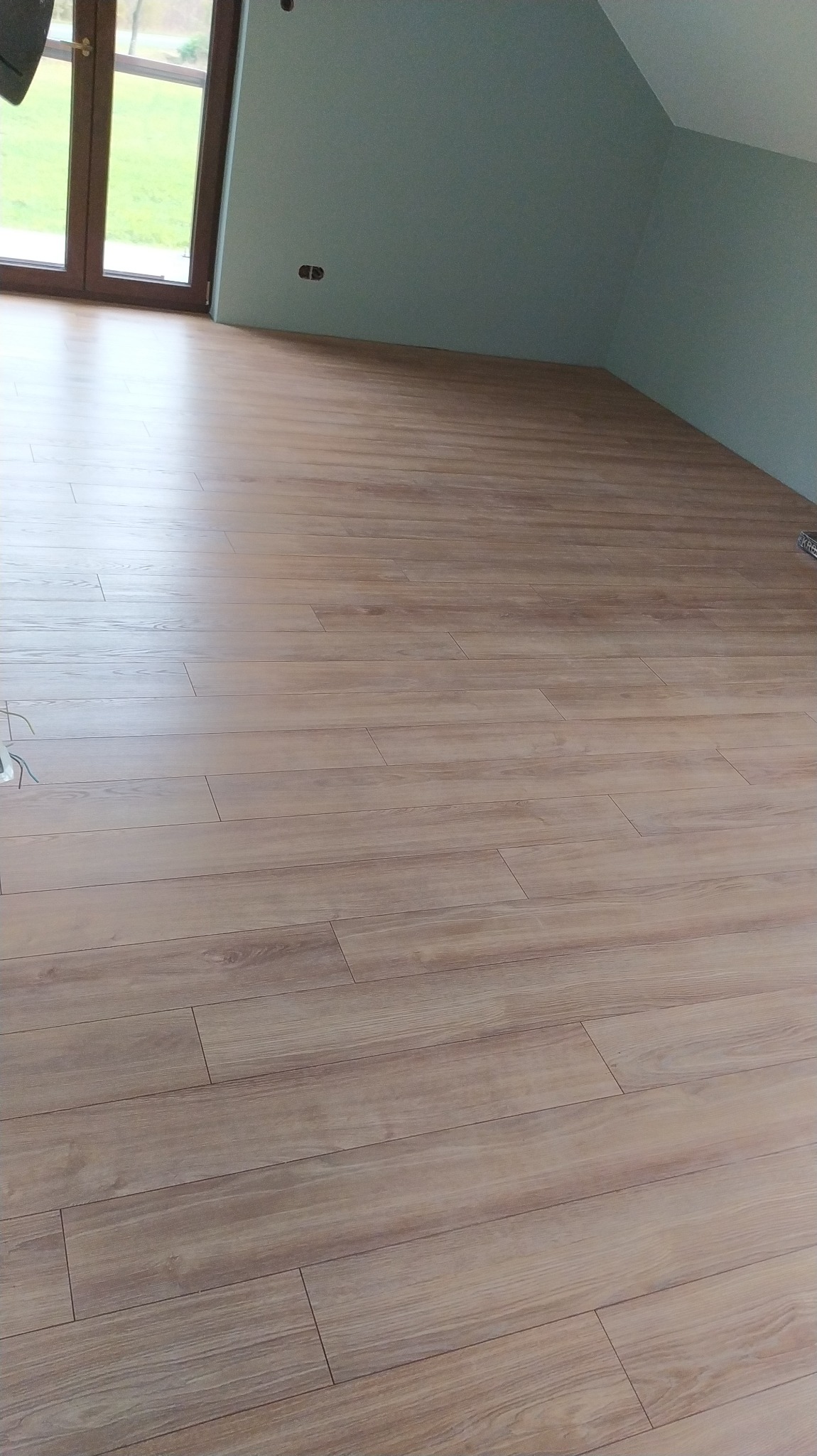 Ułożone panele podłogowe w pokoju na poddaszu, widoczne okno i fragment ściany pomalowanej na jasny błękit.