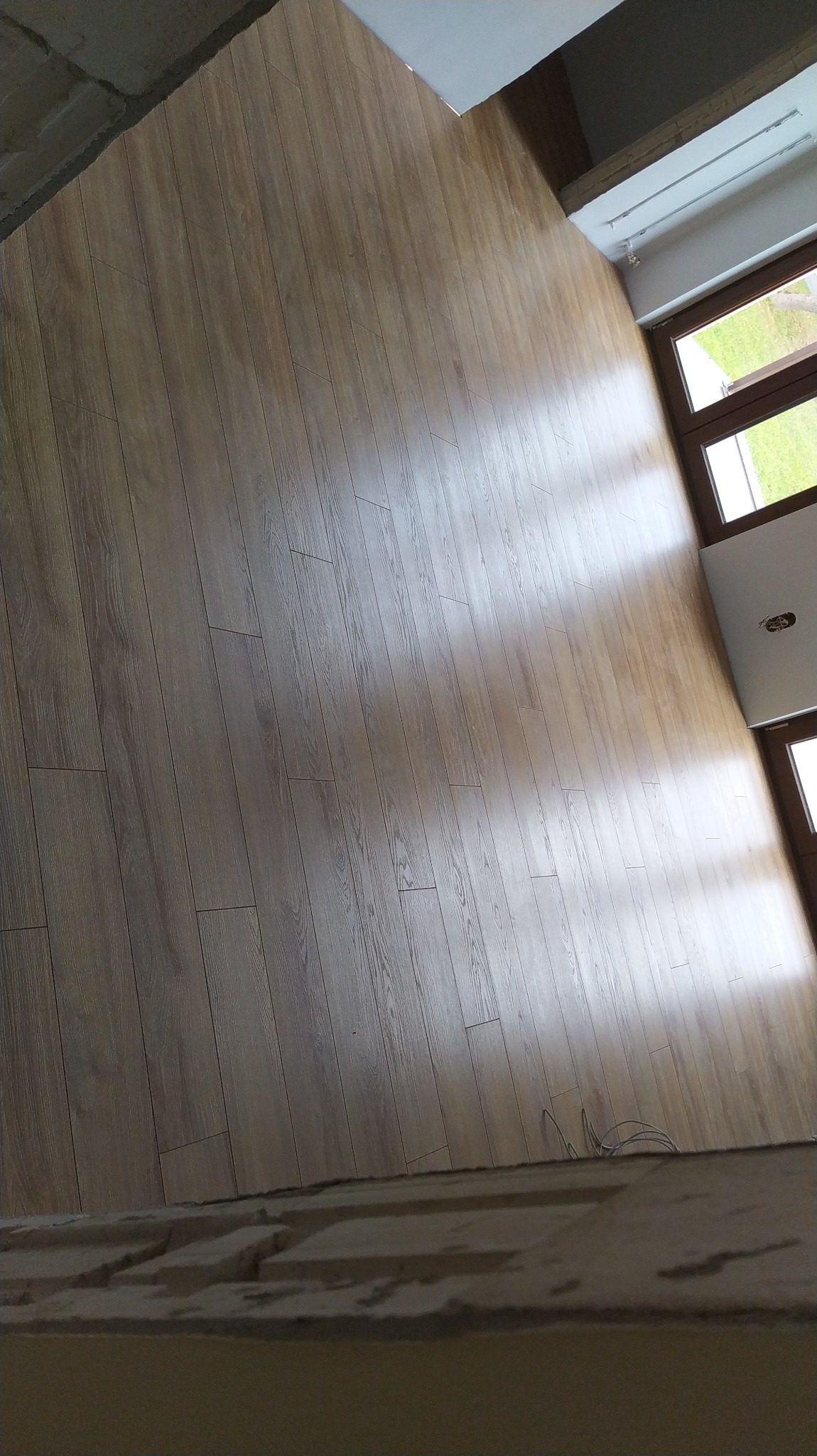 Jasnobrązowe panele ułożone w nowym pomieszczeniu, widoczne okno z zielenią za szybą i fragment surowego sufitu.