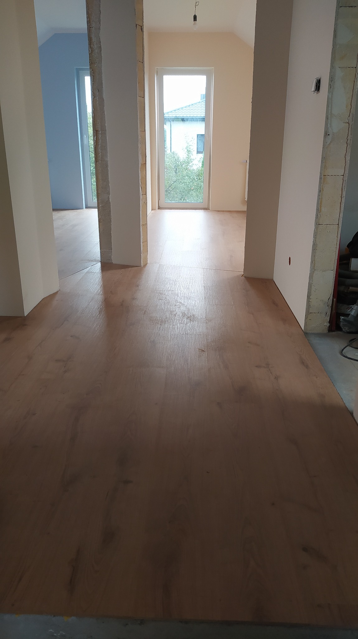 Nowo ułożona podłoga z paneli w jasnym odcieniu drewna, widok z perspektywy niskiego kąta, z fragmentami ścian w trakcie wykończenia i oknami w tle.