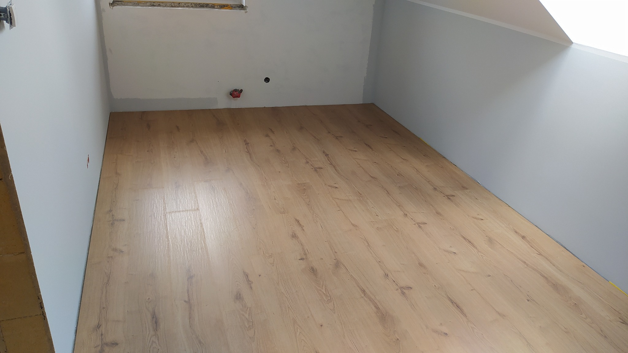 Jasne panele podłogowe ułożone w pokoju na poddaszu, widoczny fragment ściany z niedomalowanym fragmentem i otworem po gniazdku.