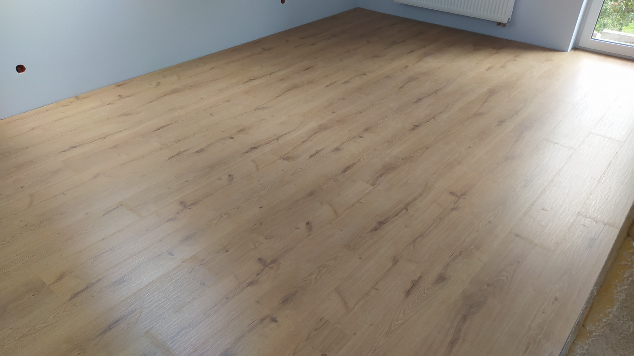 Jasne panele podłogowe imitujące drewno ułożone w pokoju, widoczne ściany w odcieniach szarości i biały grzejnik.