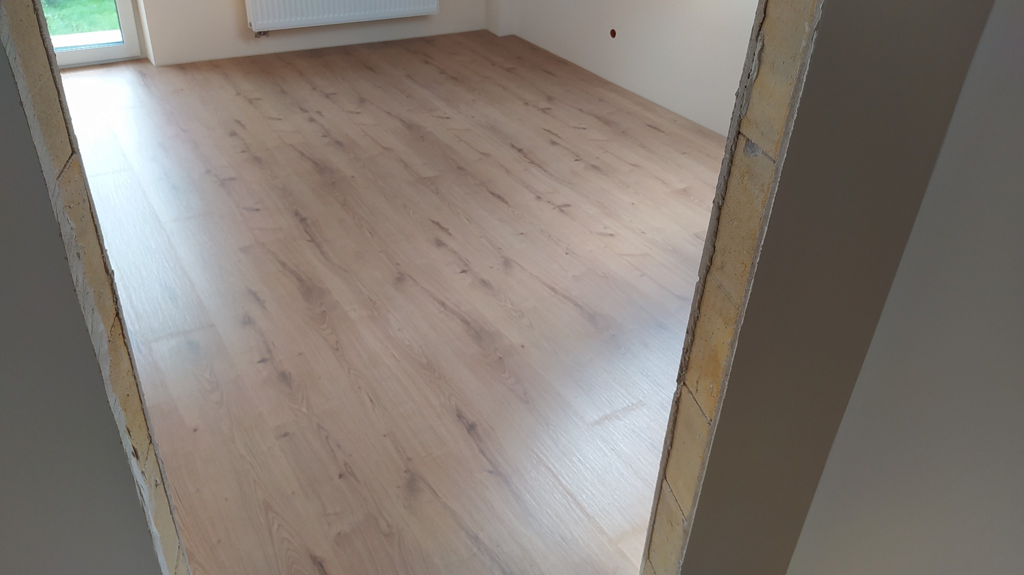 Jasne panele podłogowe ułożone w nowym pomieszczeniu, widoczne przez surową framugę drzwi.