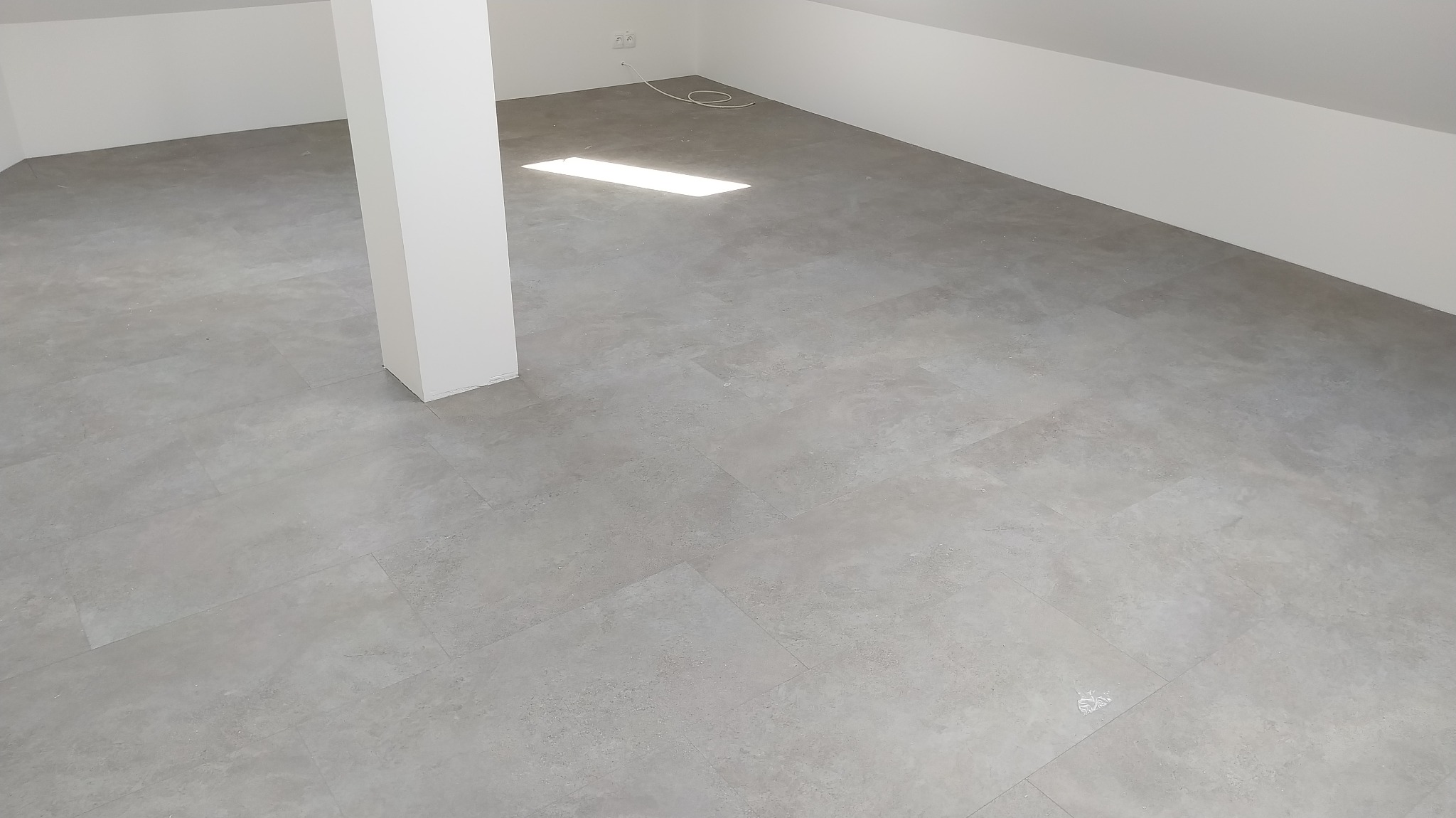 Jasnoszara podłoga z paneli w nowym, pustym pomieszczeniu pod skosem, z białym słupem konstrukcyjnym i kablem elektrycznym widocznym w rogu.