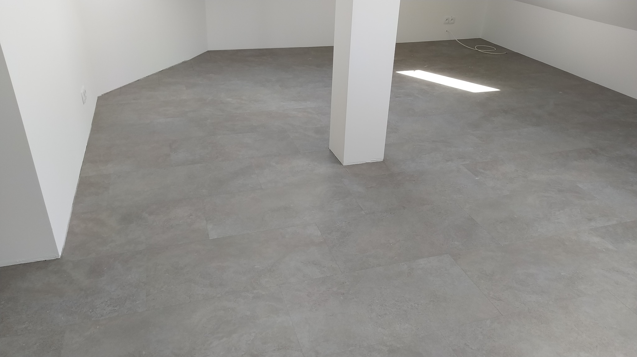 Ułożone szare panele podłogowe w jasnym, nowym pomieszczeniu z kolumną nośną i wpadającym światłem słonecznym.