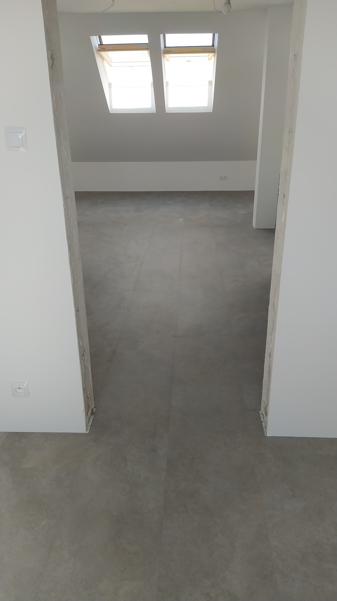 Wnętrze poddasza z dwoma oknami dachowymi i szarą, gładką podłogą z widocznymi fugami, prawdopodobnie panele lub parkiet, w stanie surowym, z widocznymi niedoskonałościami wykończenia ścian.