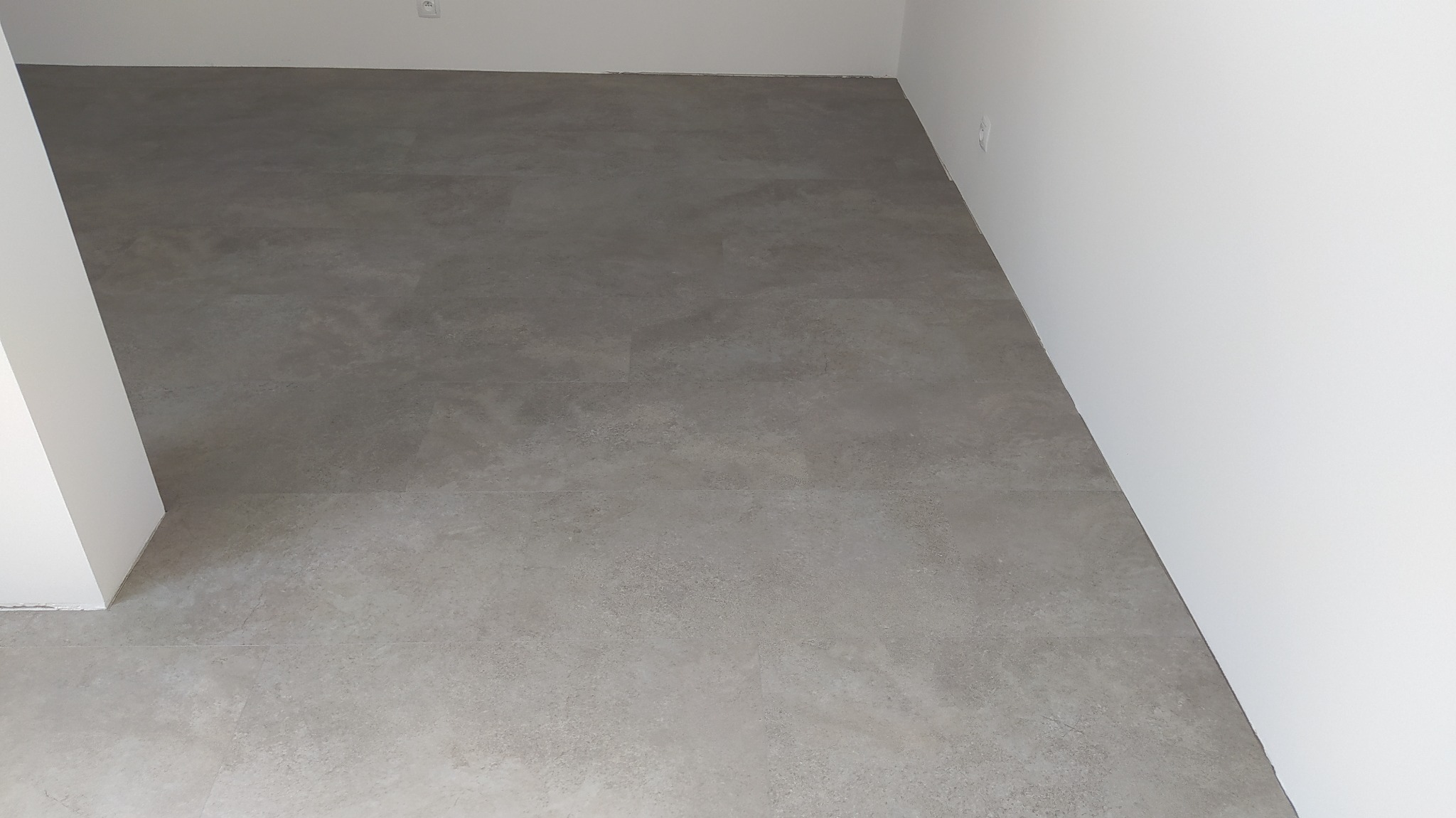 Szare panele podłogowe imitujące kamień, ułożone w nowym, jasnym pomieszczeniu z białymi ścianami i gniazdkiem elektrycznym.