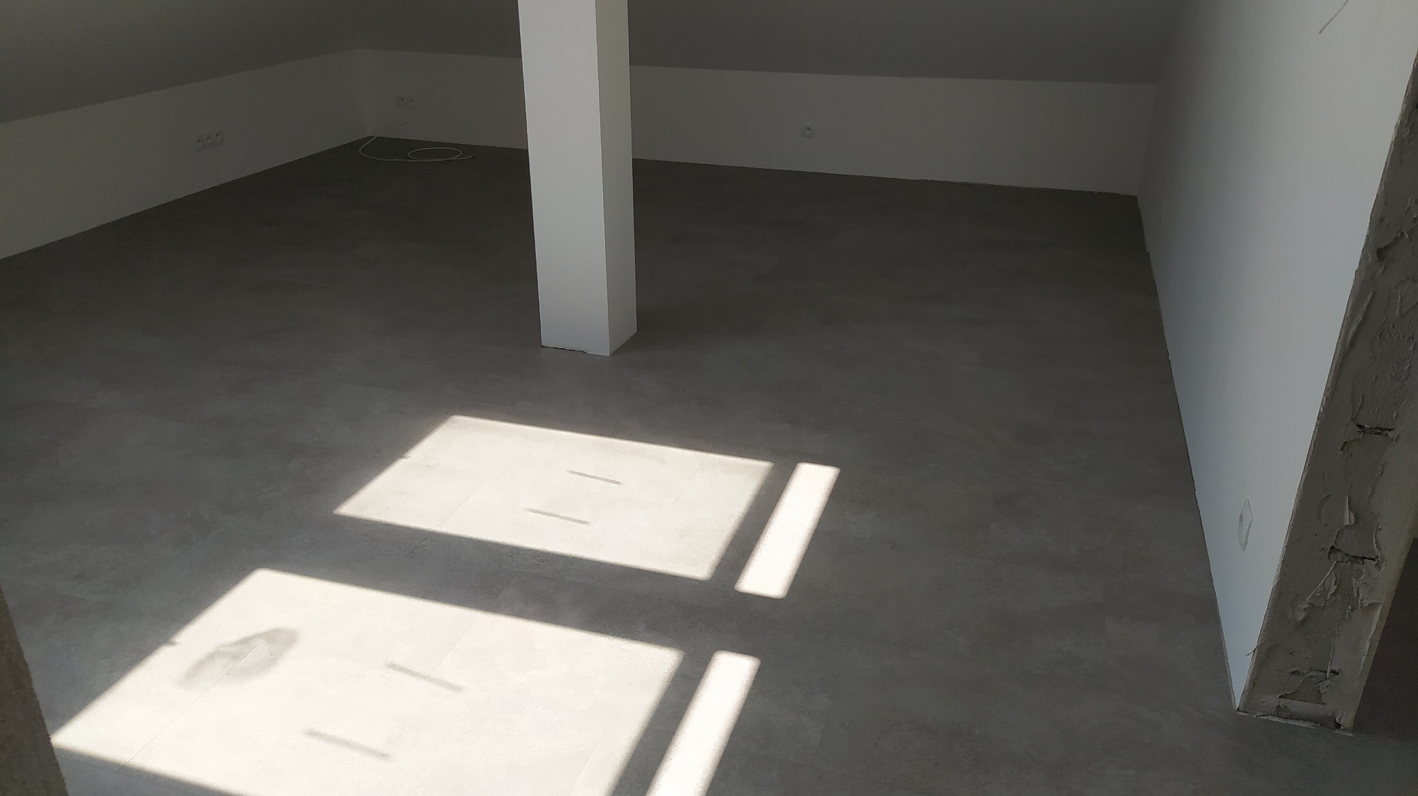 Szara, jednolita podłoga w surowym, jasnym pomieszczeniu na poddaszu z białym słupem konstrukcyjnym, oświetlona promieniami słońca wpadającymi przez okno, tworzącymi geometryczne wzory.