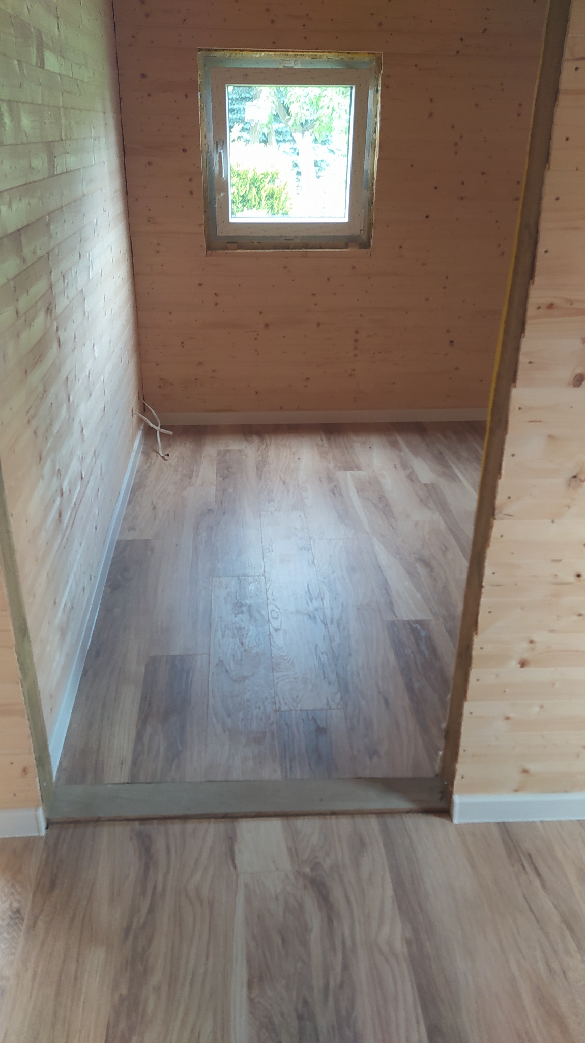 Wnętrze pomieszczenia wykończonego drewnianą boazerią i panelami podłogowymi imitującymi drewno, z oknem w tle, widok z progu.