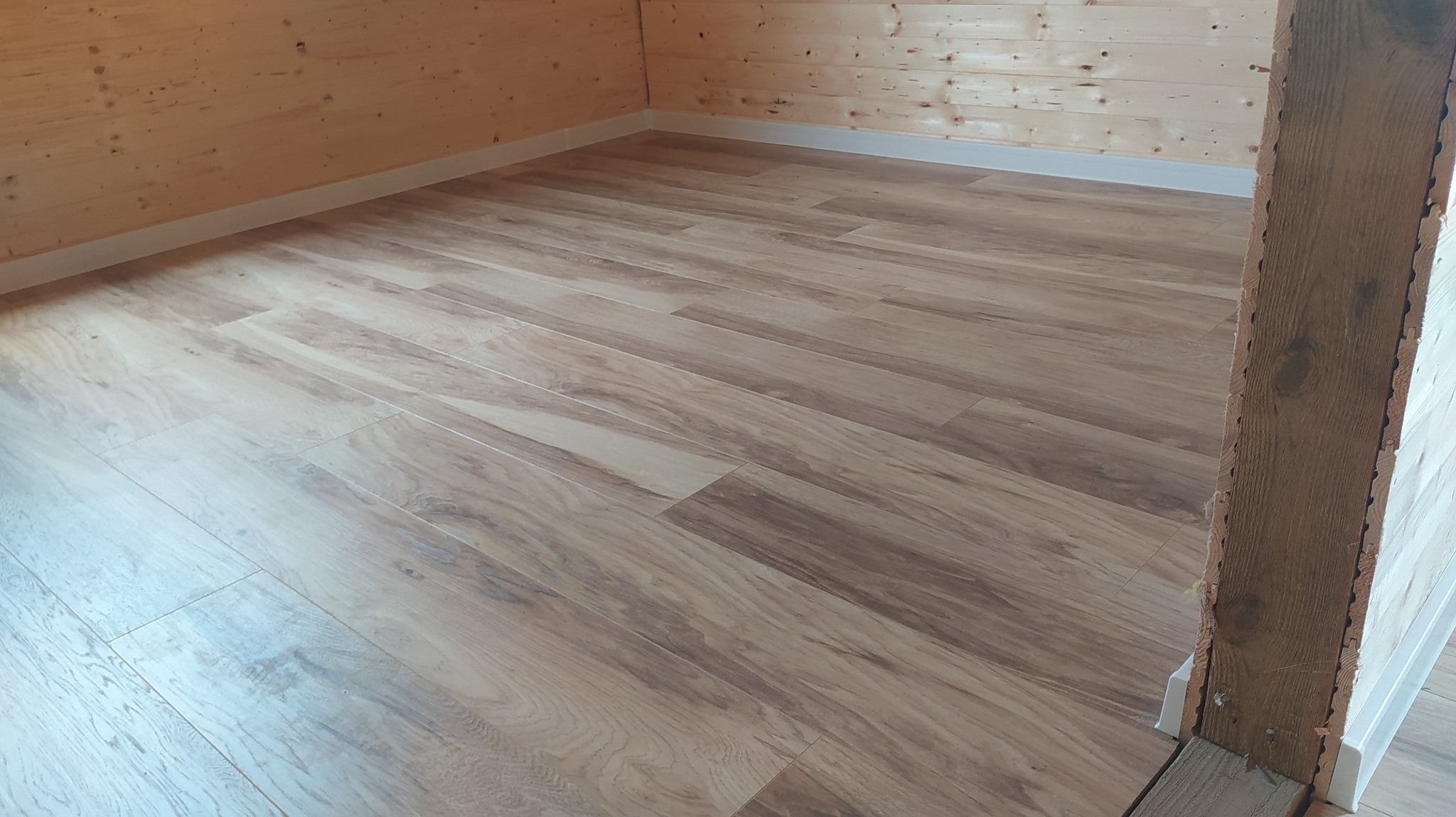 Jasne panele podłogowe imitujące drewno, ułożone w nowym pomieszczeniu z boazerią, widoczny fragment nieukończonej ściany po prawej.