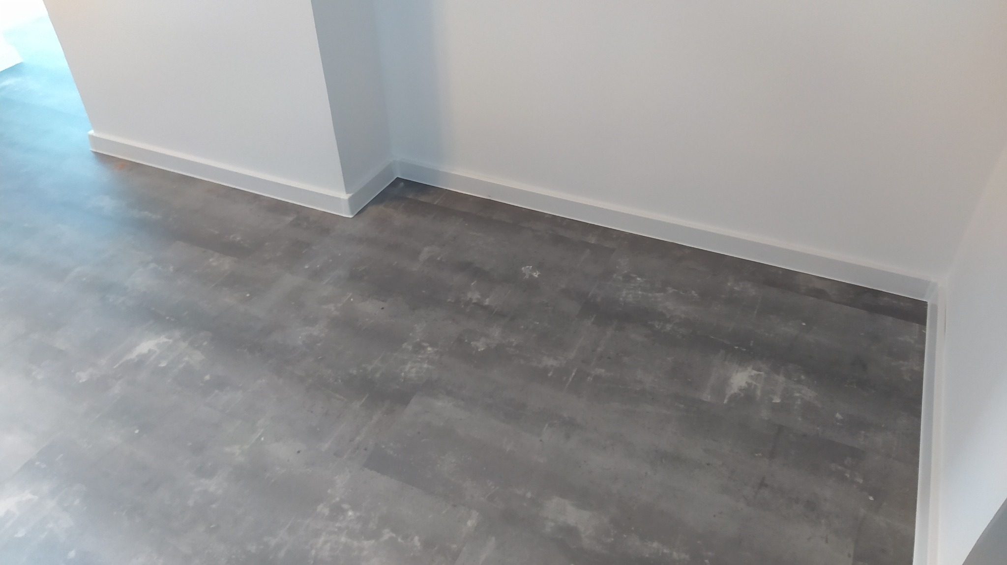 Szare panele podłogowe ułożone w rogu pokoju z białymi listwami przypodłogowymi.