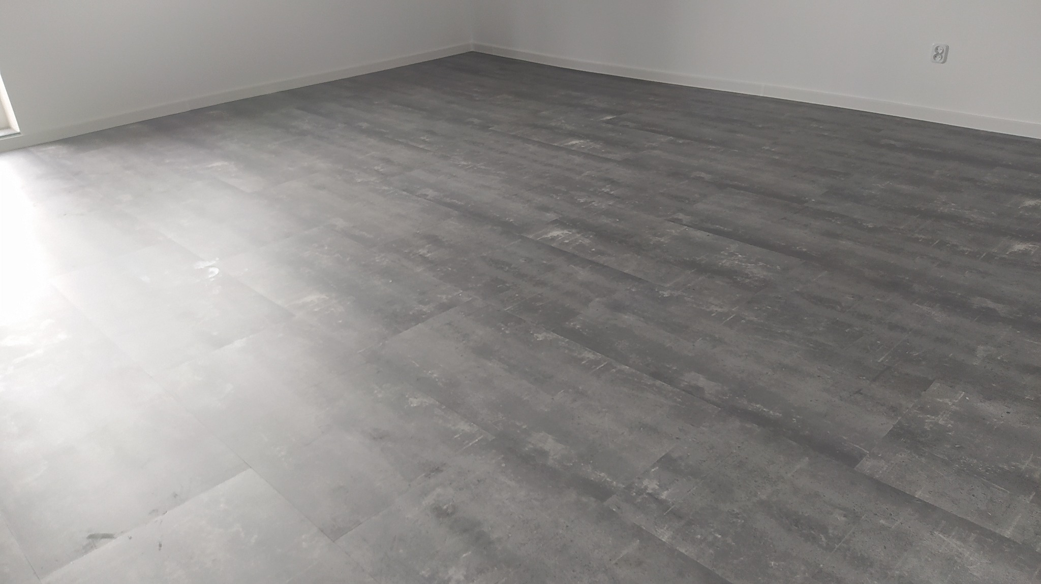 Szara podłoga z paneli imitujących beton w pustym, białym pomieszczeniu, widoczny fragment listwy przypodłogowej i gniazdko elektryczne.