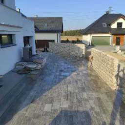Nowoczesna posesja z szarą kostką brukową ułożoną na dużej powierzchni, obok domu i ogrodzenia gabionowego, w tle widoczne inne budynki.