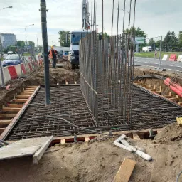 Zbrojenie fundamentu z widocznym szalunkiem drewnianym i pracownikiem w oddali na tle miejskiego krajobrazu.