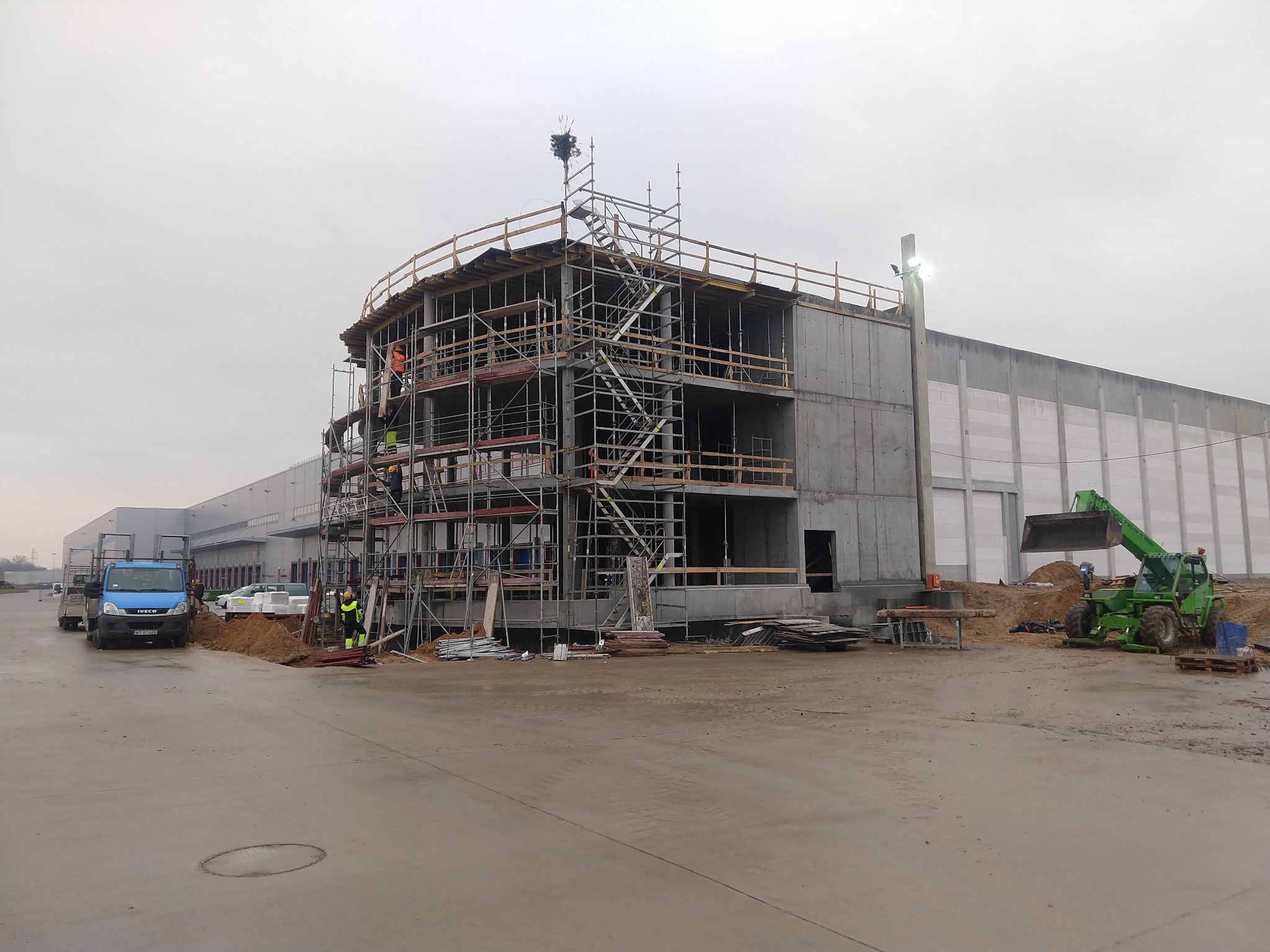 Budowa hali magazynowej z betonu, widoczne rusztowania, pracownicy w odblaskowych ubraniach, w tle sprzęt budowlany i ciężarówki.