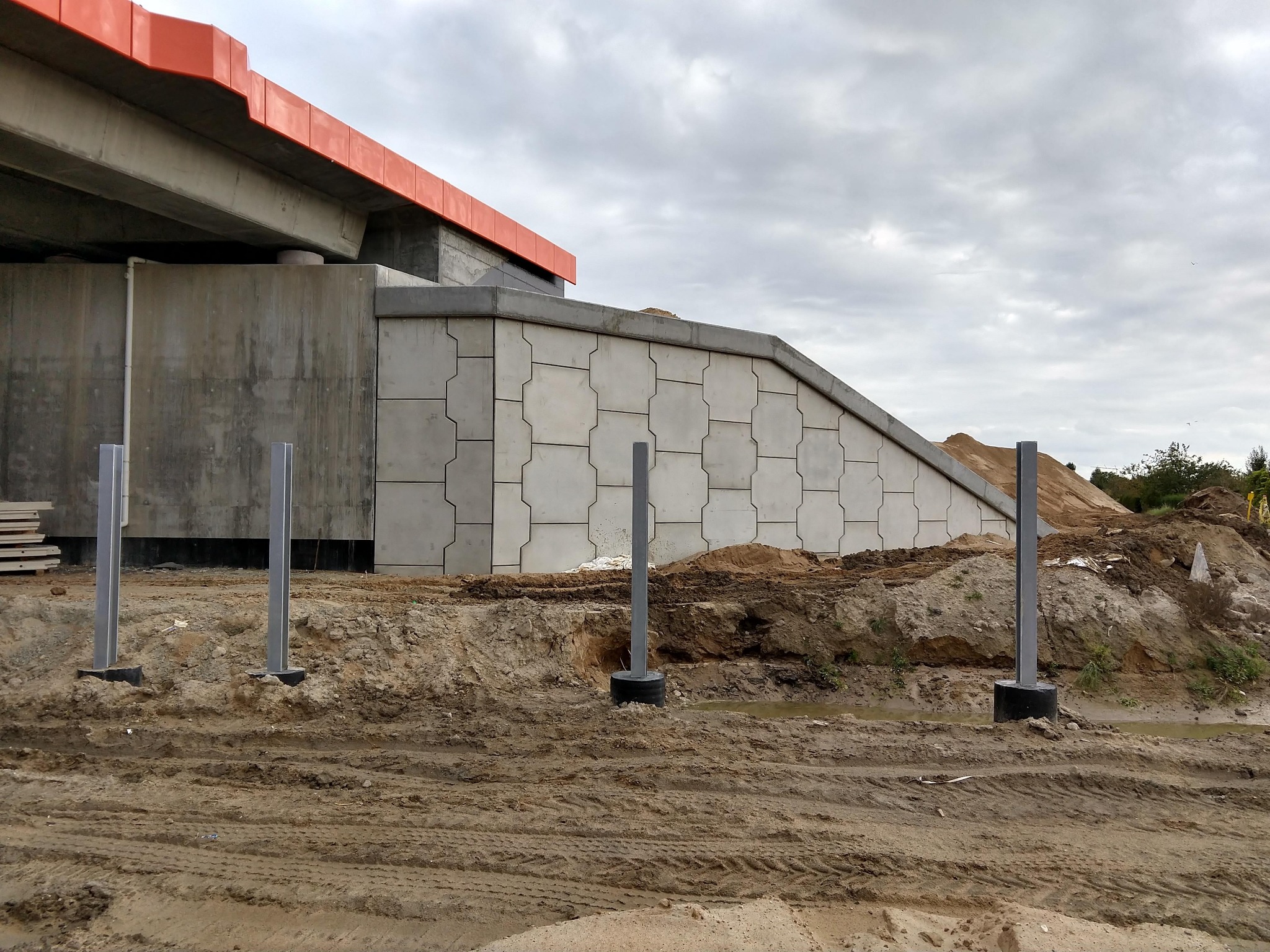 Betonowa konstrukcja mostu z czerwonymi elementami wykończeniowymi, widoczne metalowe słupki i teren budowy z ziemią i śladami opon.